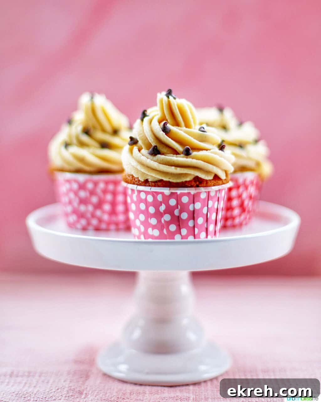 Chocolate Chip Cupcakes with Cookie Dough Frosting Recipe 2 Three Chocolate Chip Cupcakes with Cookie Dough Frosting on a white cake pedestal with pink background
