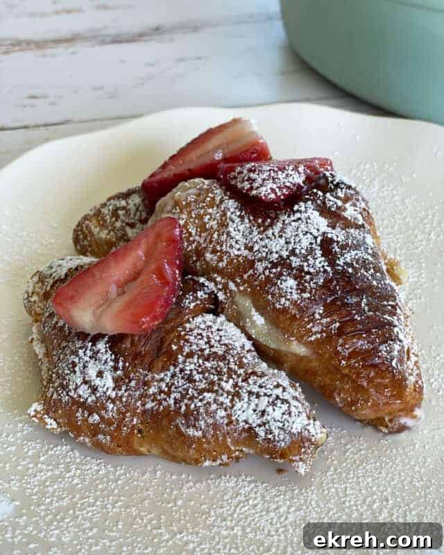 two strawberry cheesecake french toast croissants on a plate topped with powdered sugar and sliced strawberries