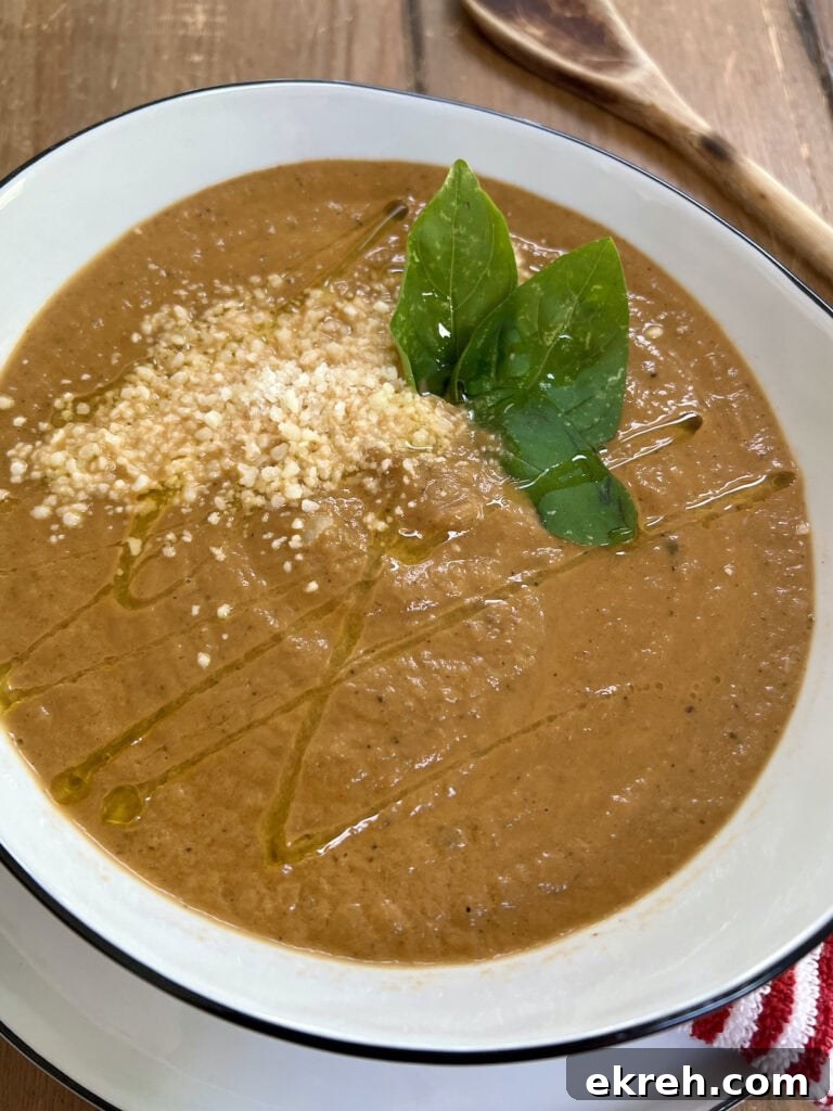 Creamy Eggplant Soup in white soup bowl with basil leaf, olive oil and grated cheese garnish, with wooden spoon in background.