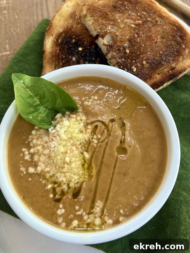 Creamy Eggplant Soup in white ramekin with basil leaf, olive oil and grated cheese garnish, over green towel, with toast in background.