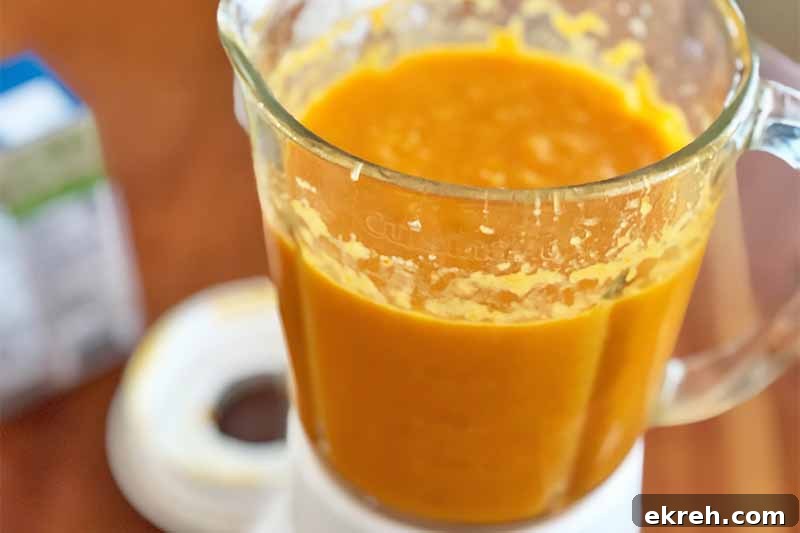 Dukkah-Spiced Roasted Carrot and Onion Soup Recipe 9 Horizontal image of a pureed orange mixture in a blender on a wooden table.