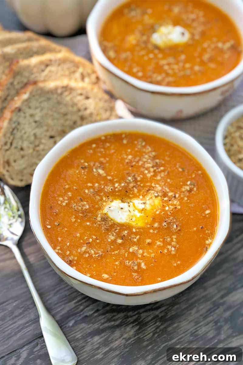 Dukkah-Spiced Roasted Carrot and Onion Soup Recipe 4 Vertical image of two bowls filled with a bright orange puree garnished with a spice blend and a dollop of yogurt in the middle, next to a metal spoon and slices of bread.