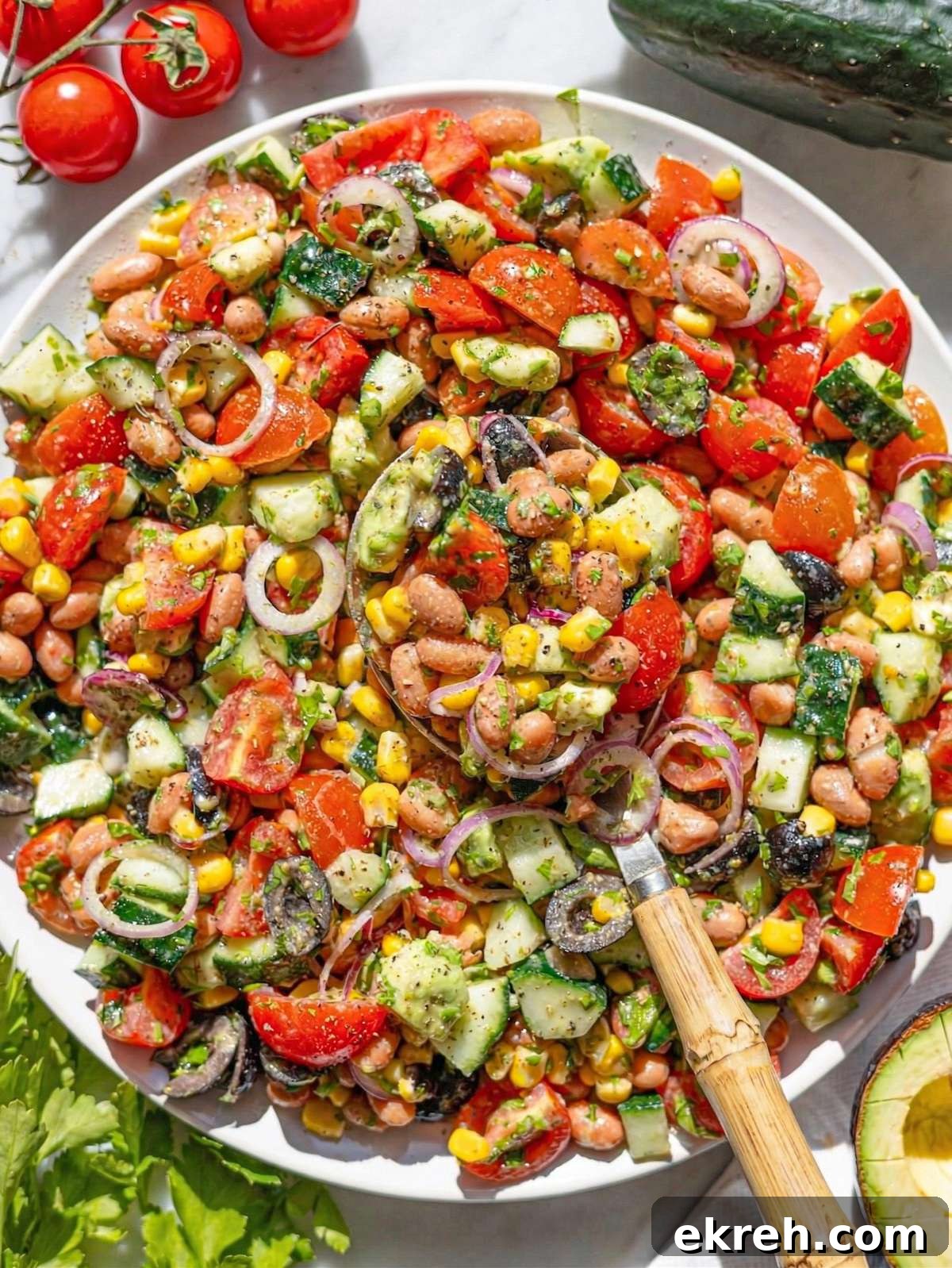 Pinto bean salad in bowl on marble table with avocado, tomatoes, cucumber, and herbs.