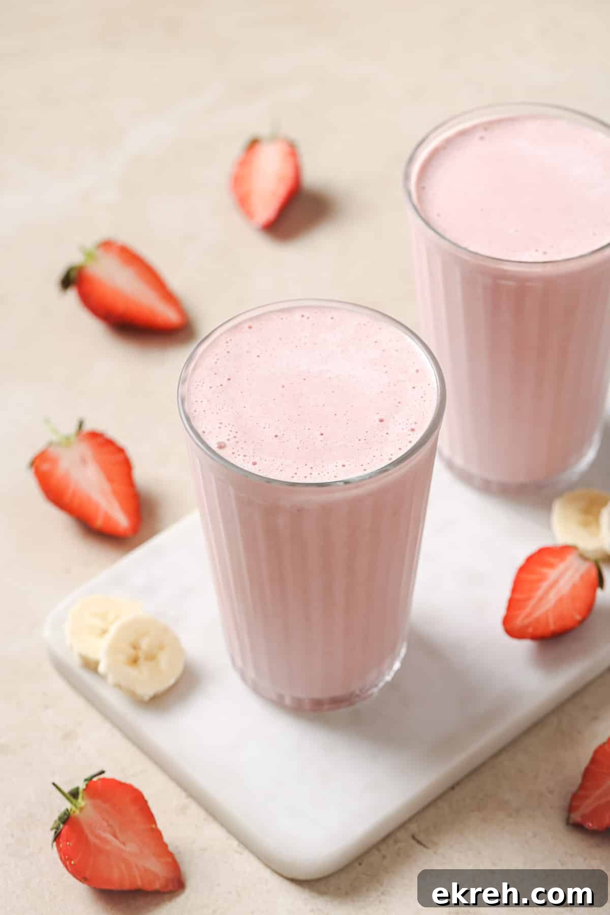 Two glasses of pink strawberry banana smoothie on a white rectangular tray, surrounded by sliced strawberries and banana pieces on a beige surface.
