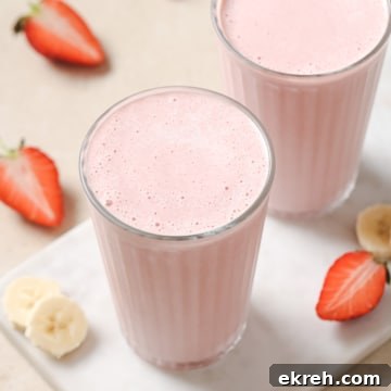 Two glasses of pink strawberry banana smoothie on a white surface, with sliced strawberries and bananas around.