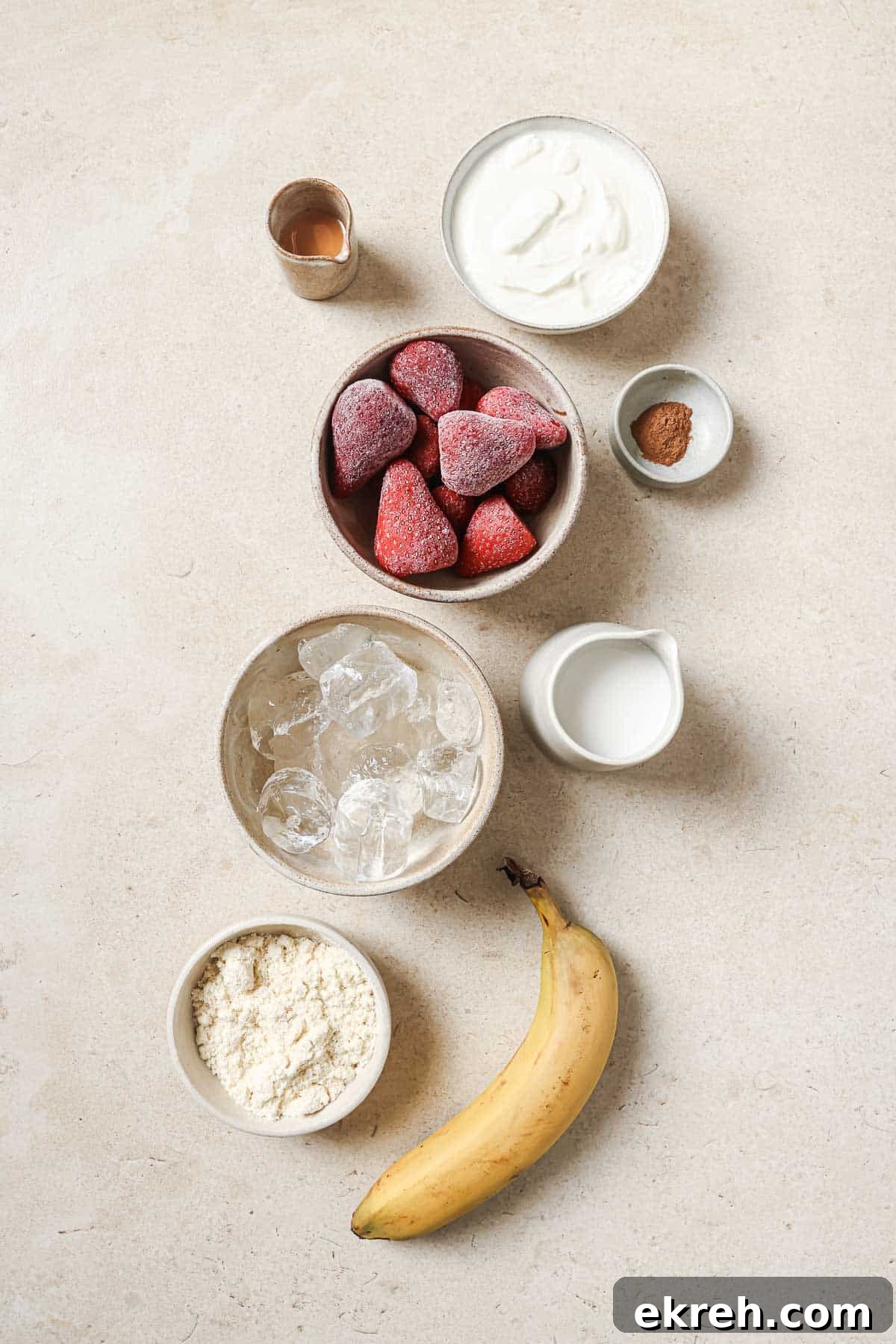 Ingredients for a smoothie: a banana, frozen strawberries, ice cubes, yogurt, protein powder, cinnamon, milk, and a small amount of liquid in a shot glass. All items are placed neatly on a light countertop.