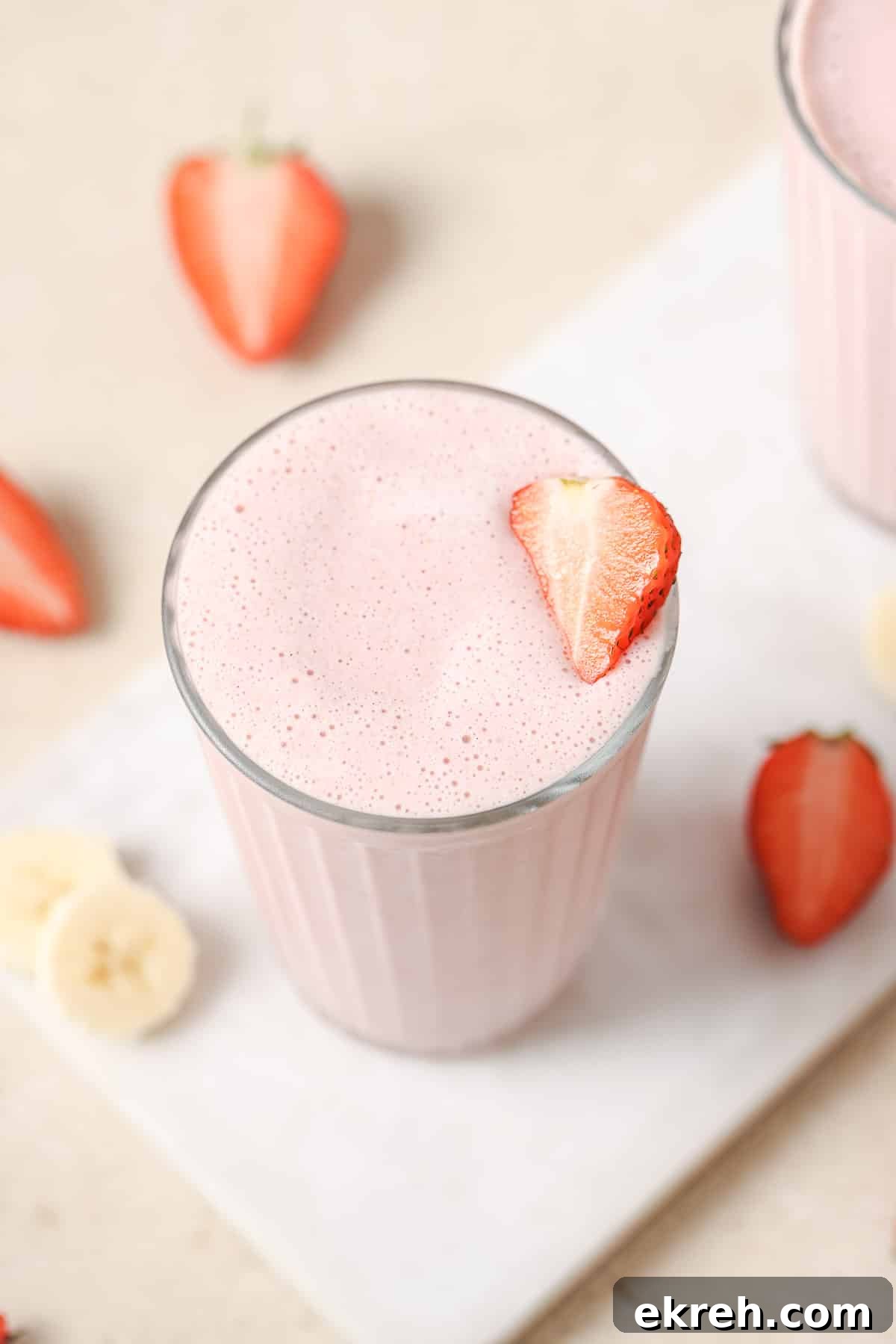 A glass of strawberry smoothie garnished with a slice of strawberry, surrounded by additional strawberry pieces and banana slices on a white surface.