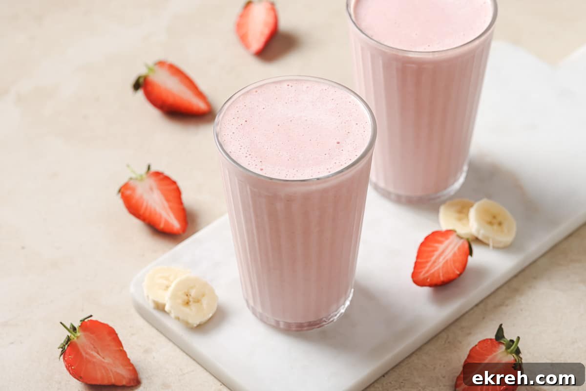 Two glasses of strawberry banana smoothie are placed on a white marble surface, surrounded by sliced strawberries and banana pieces.