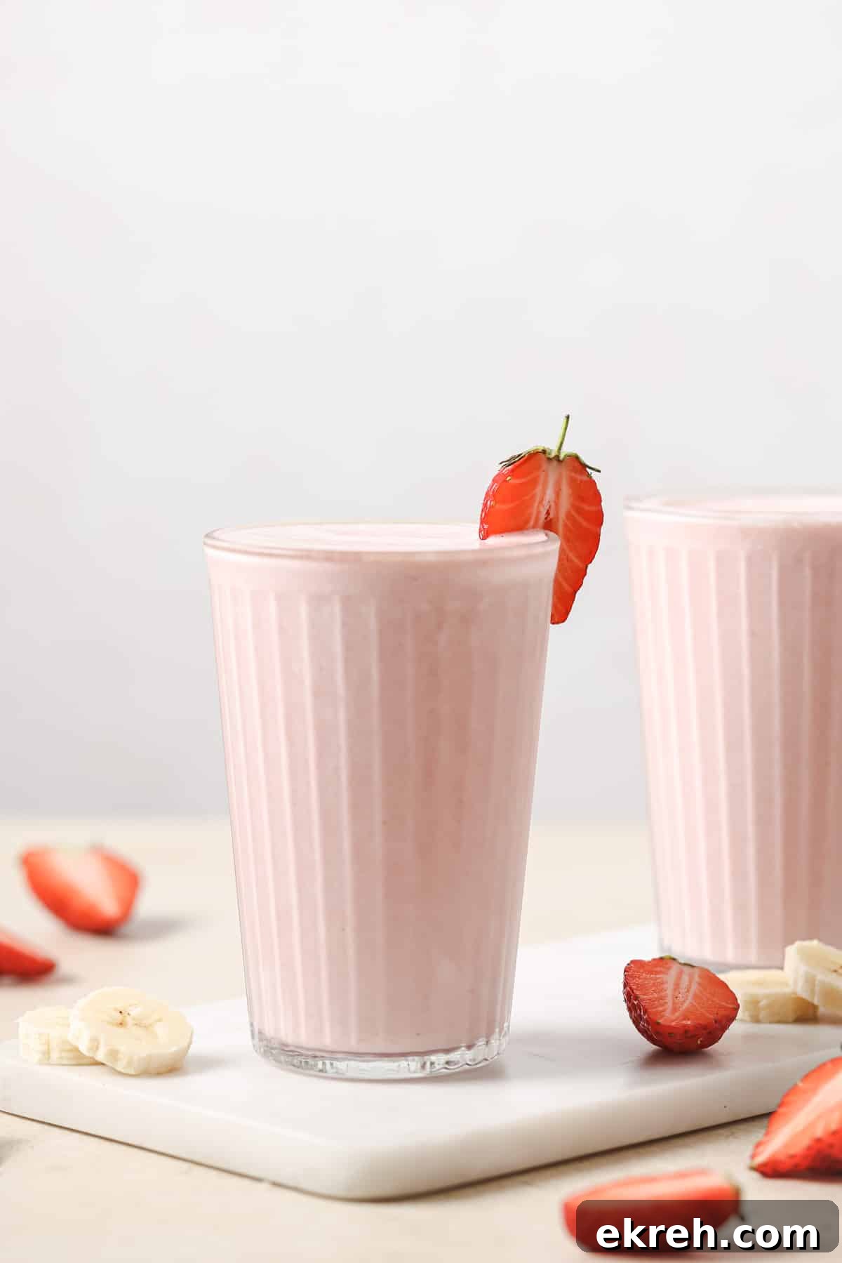 A glass of pink smoothie garnished with a strawberry slice, placed on a white surface. Sliced strawberries and banana pieces are scattered around. Another glass is partly visible in the background.