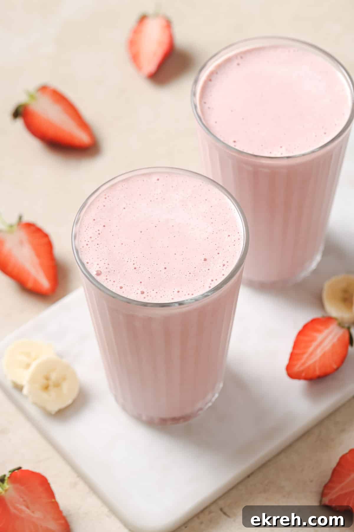 Two glasses of strawberry banana smoothie on a white countertop, surrounded by strawberry halves and banana slices.