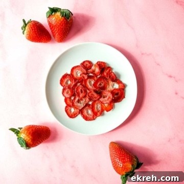 A white plate of air fryer dehydrated strawberries sits on a pink marble background surrounded by fresh strawberries.