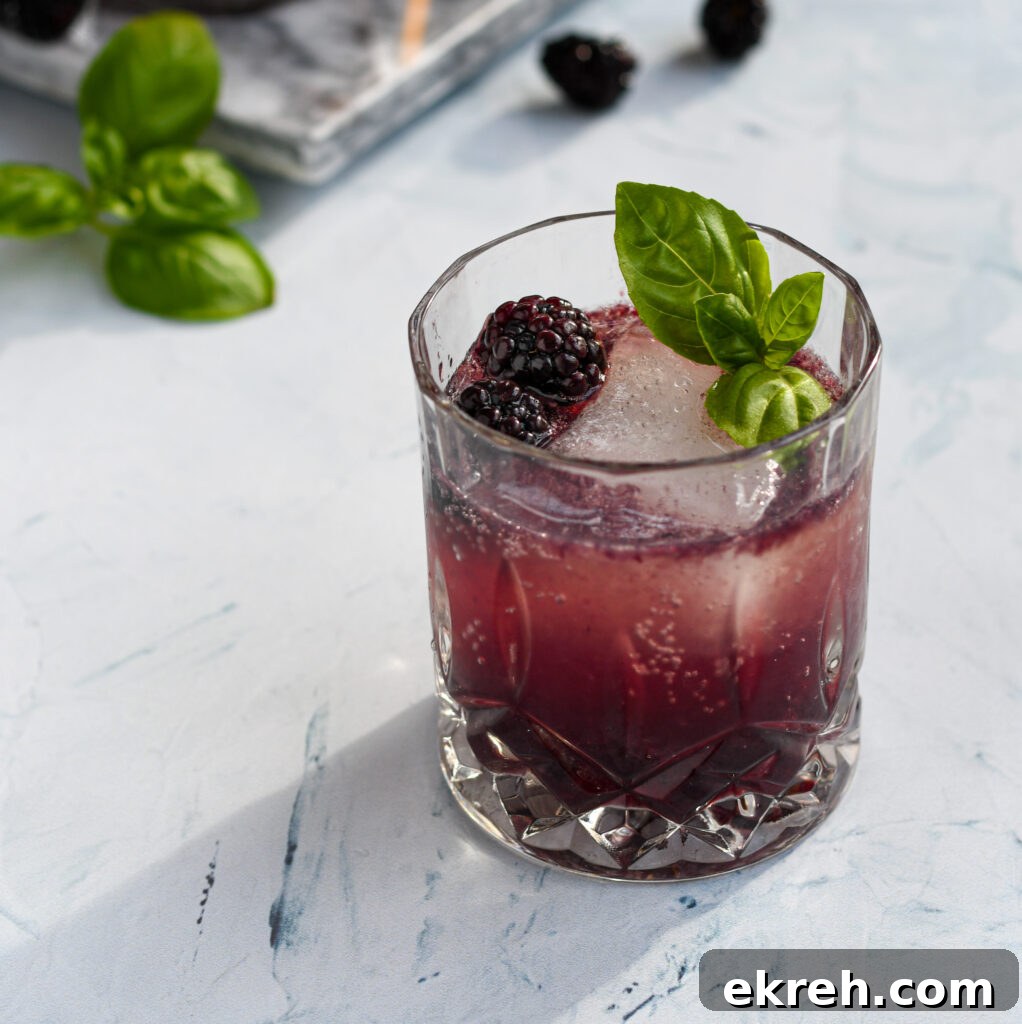 an old fashioned glass filled with a purple blackberry bourbon smash garnished with a large ice cube, blackberries, and basil