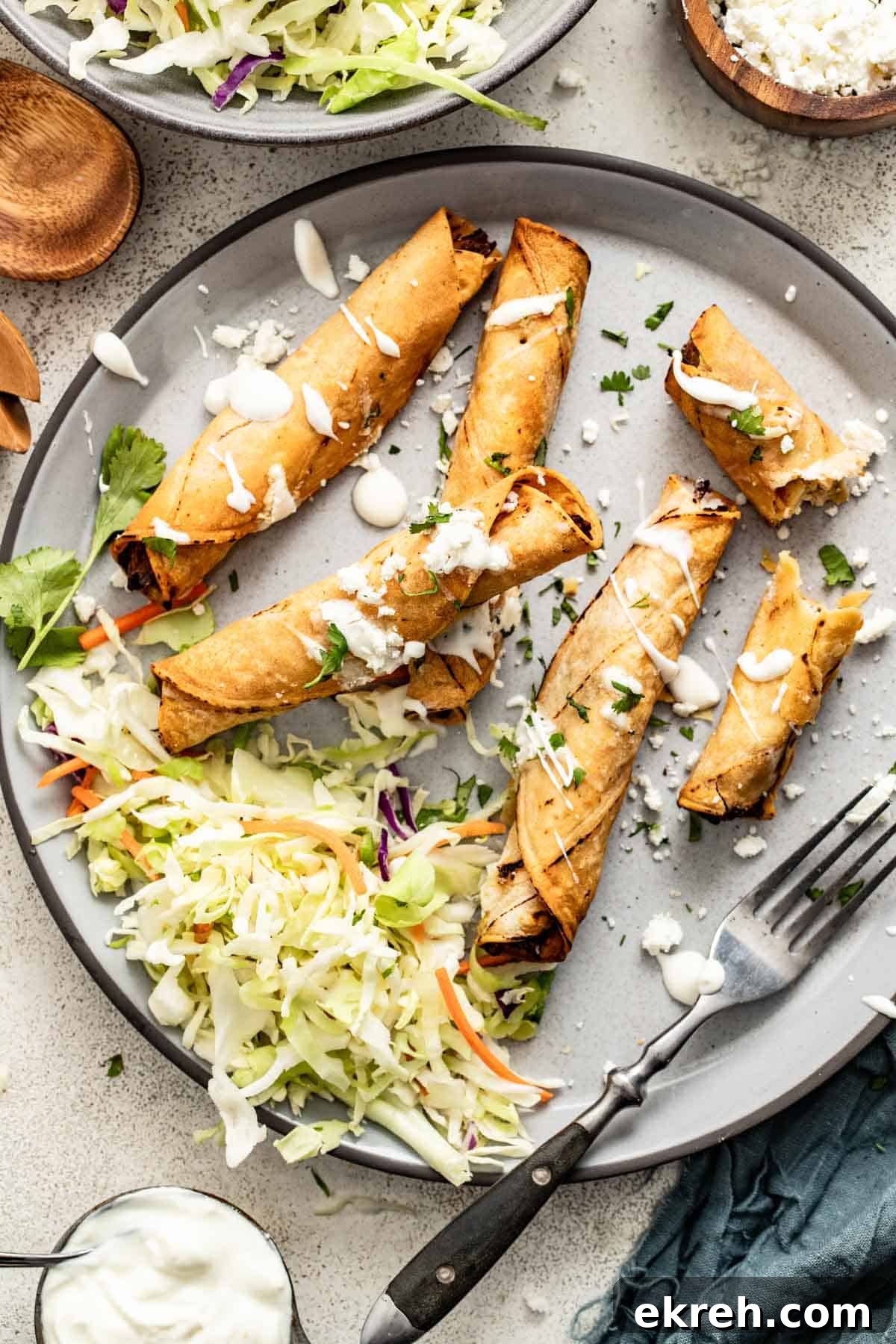 How to Make Crispy Air Fryer Chicken Taquitos 9 Chicken taquitos stacked next to shredded slaw, a whipped cottage cheese, and cilantro.