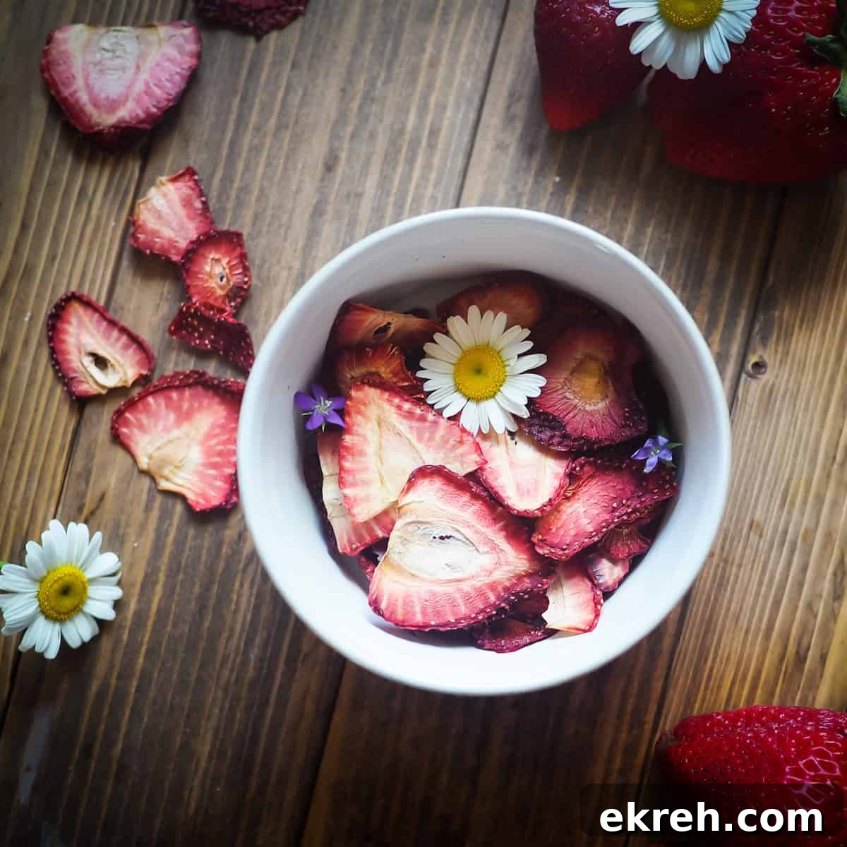 A bowl of dehydrated strawberries on a wood background with some daisies and small purple flowers scattered around.