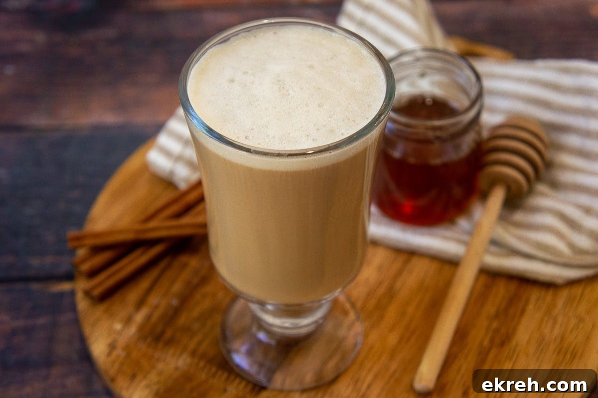 Creamy Oat Milk Honey Latte Recipe 2 a glass coffee cup with a creamy brown latte beside a small honey pot and cinnamon sticks.
