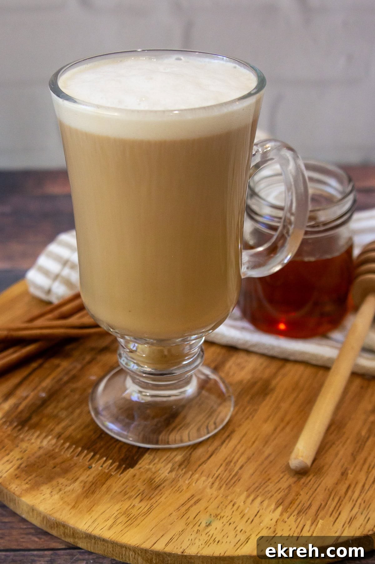 Creamy Oat Milk Honey Latte Recipe 6 a glass coffee cup with a creamy brown latte beside a small honey pot and cinnamon sticks.