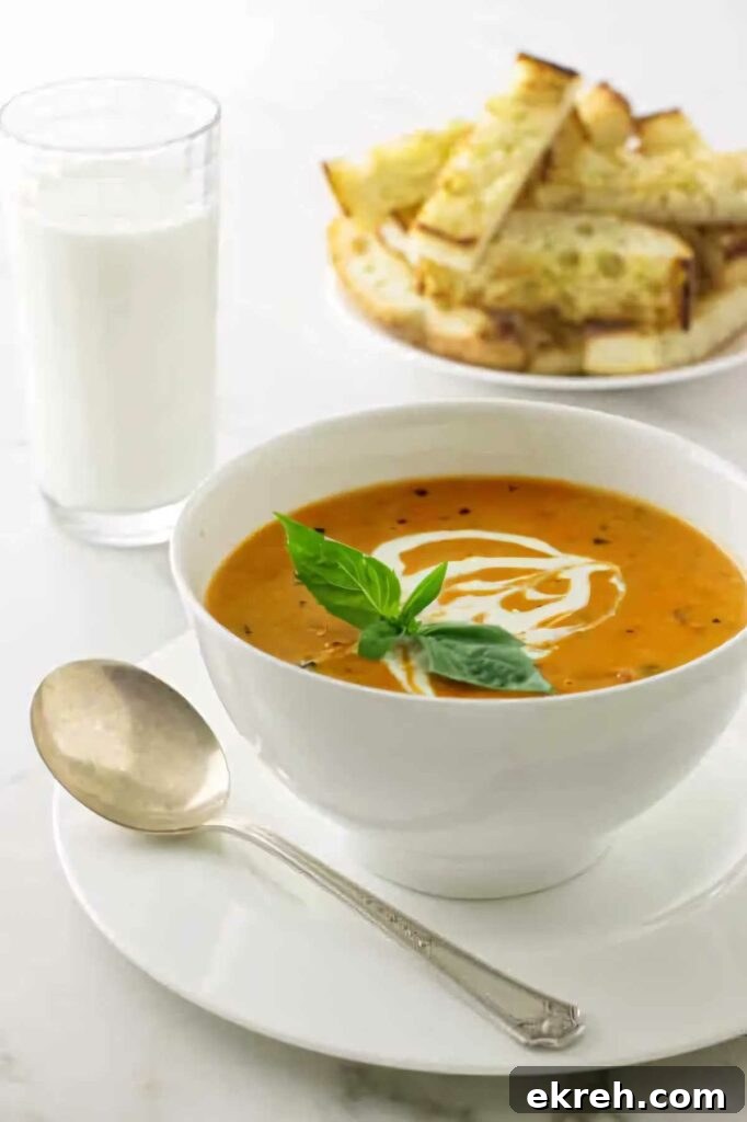 A bowl of roasted tomato soup in front of a glass of milk and some bread.