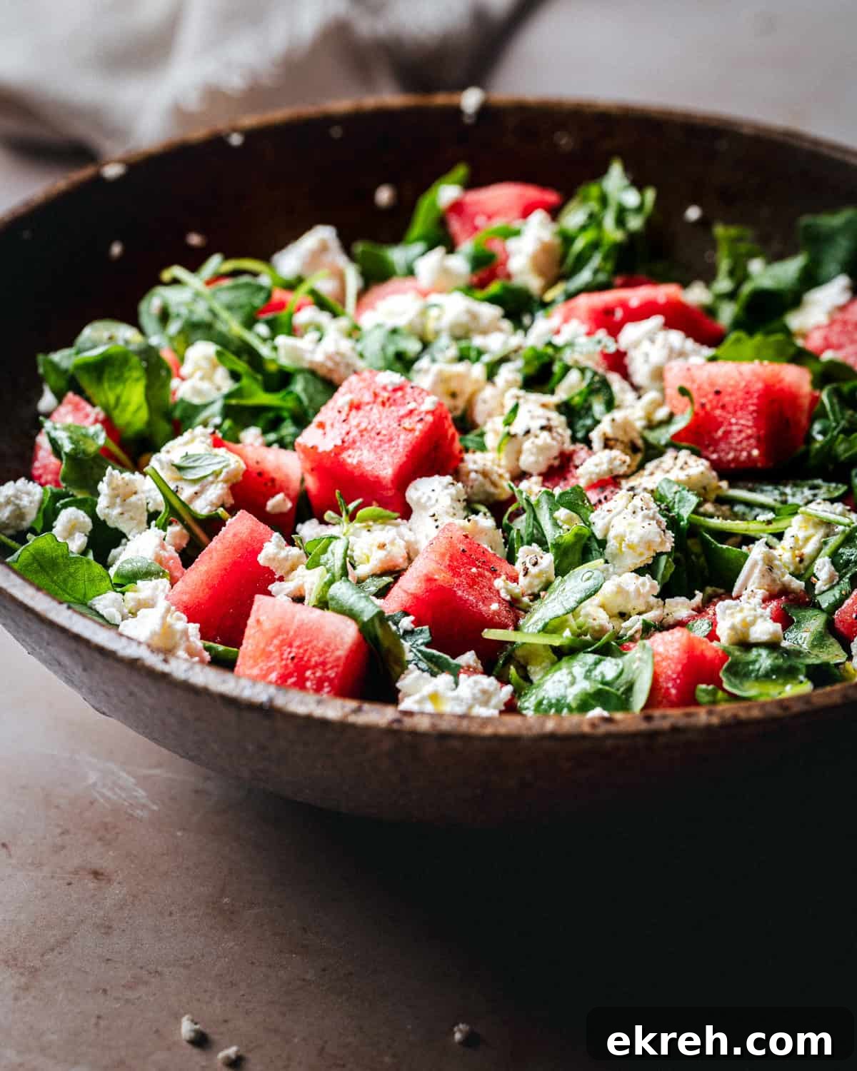 A close-up of a salad in a rustic brown bowl, featuring chunks of watermelon, crumbled feta cheese, and fresh green leaves, lightly seasoned with black pepper.