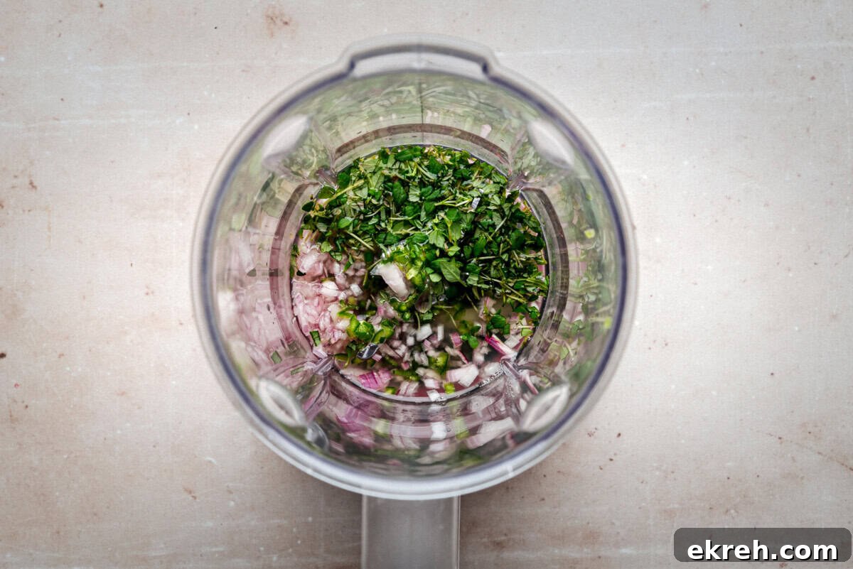 Chopped fresh herbs and diced onions inside a blender viewed from above, ready to be blended on a light-colored countertop.