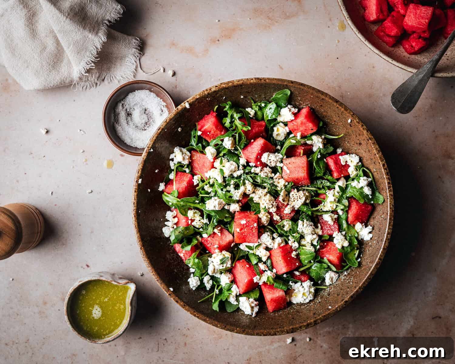 A bowl of watermelon salad with arugula and crumbled feta cheese on a light surface, surrounded by a jar of olive oil, a wooden pepper grinder, a bowl of salt, and a linen napkin.