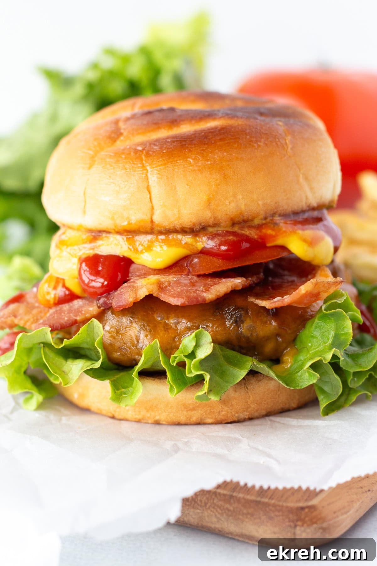 Juicy Grilled Bacon Cheeseburgers with Beef and Pork Patties 2 A cheeseburger with lettuce, bacon, cheddar cheese, ketchup, and a toasted bun is placed on parchment paper, with blurred lettuce and a tomato in the background.