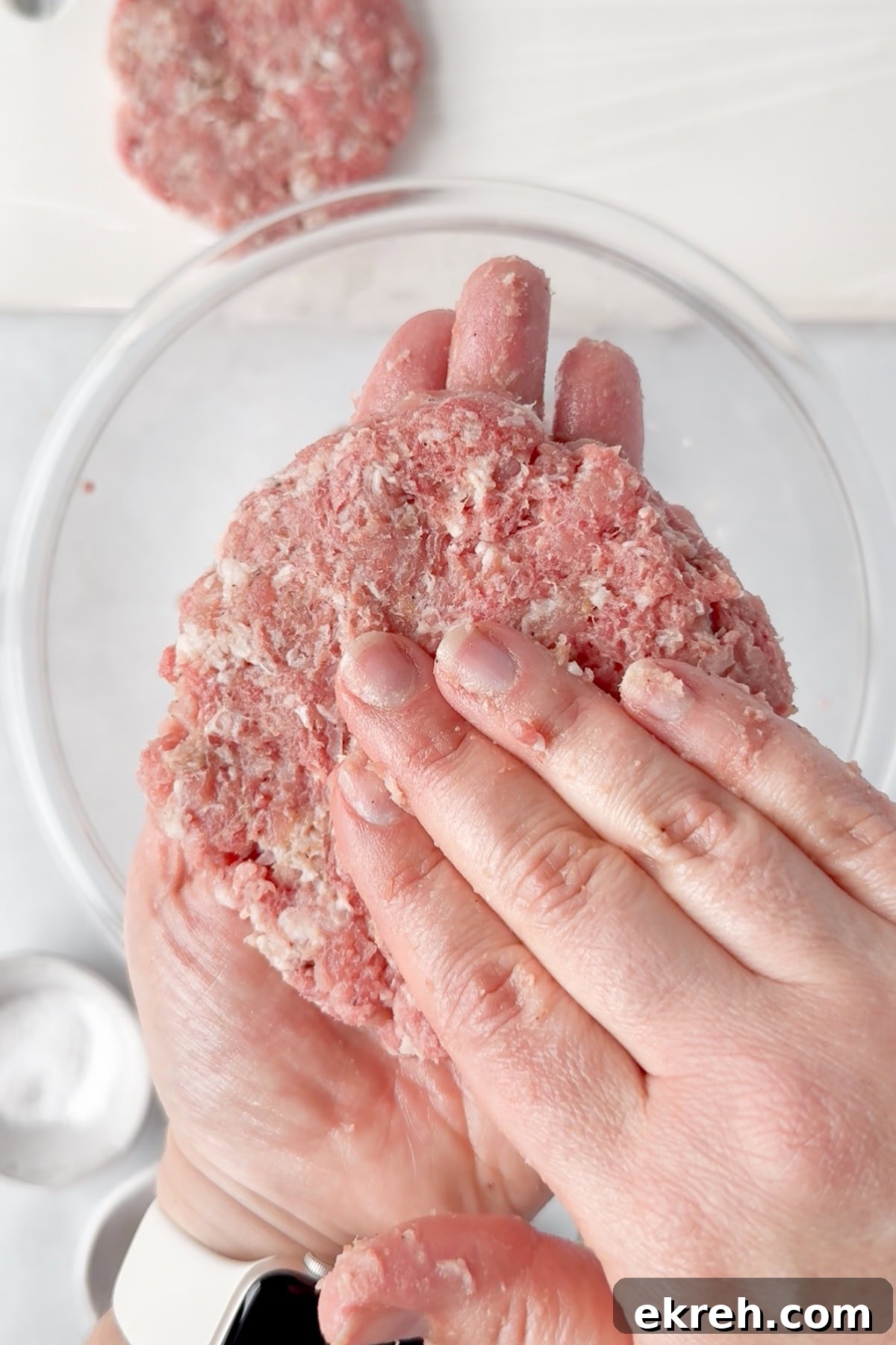 Juicy Grilled Bacon Cheeseburgers with Beef and Pork Patties 11 A close-up of hands shaping raw ground meat into a patty over a clear glass bowl, with a cutting board and another patty in the background.