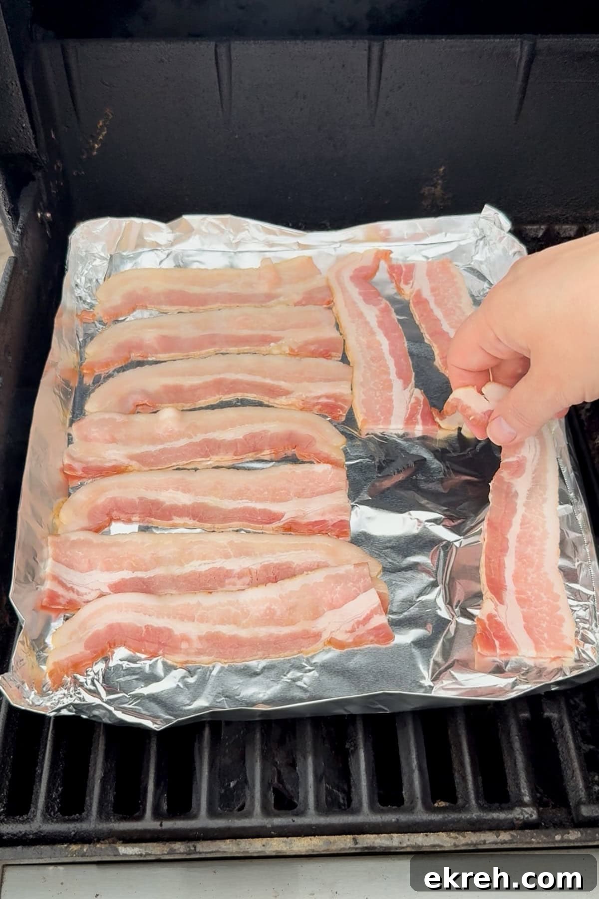 Juicy Grilled Bacon Cheeseburgers with Beef and Pork Patties 12 A hand places strips of raw bacon on a foil-lined grill grate, with several slices already arranged side by side for cooking.