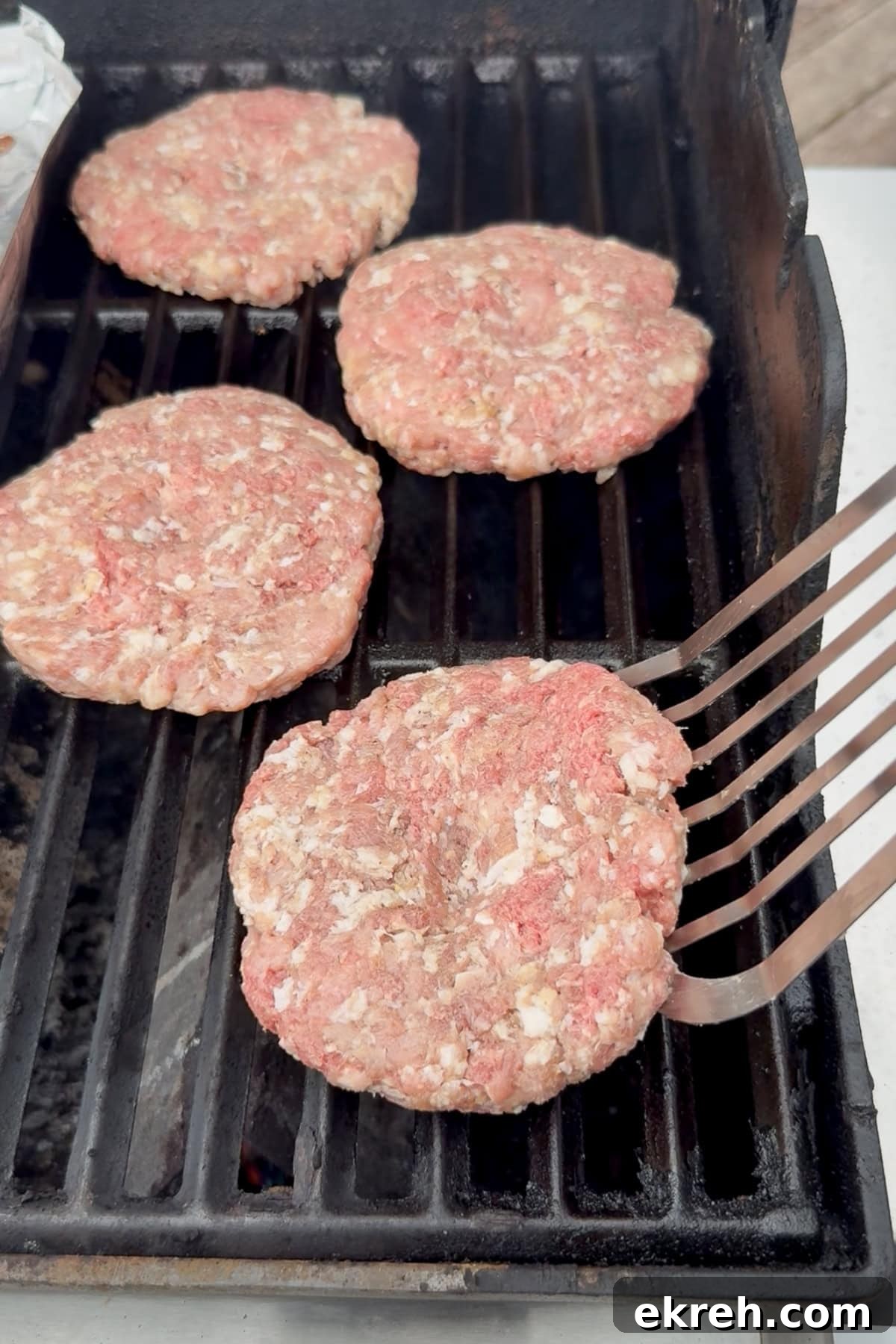 Juicy Grilled Bacon Cheeseburgers with Beef and Pork Patties 13 Four raw hamburger patties are on a grill, with one patty being lifted off a metal spatula. The grill grates are visible, and the scene appears to be outdoors.