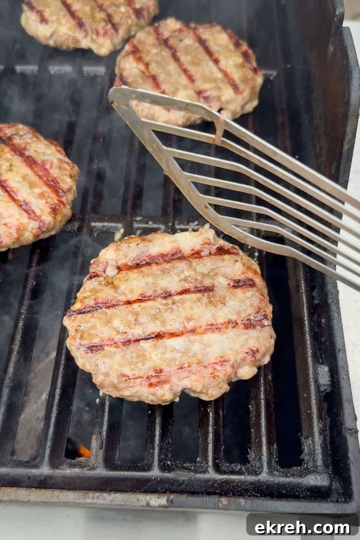 Juicy Grilled Bacon Cheeseburgers with Beef and Pork Patties 14 Three hamburger patties are being grilled, with one being flipped by a metal spatula. The patties have visible grill marks and are cooking on a black grill grate. Smoke is rising from the grill.