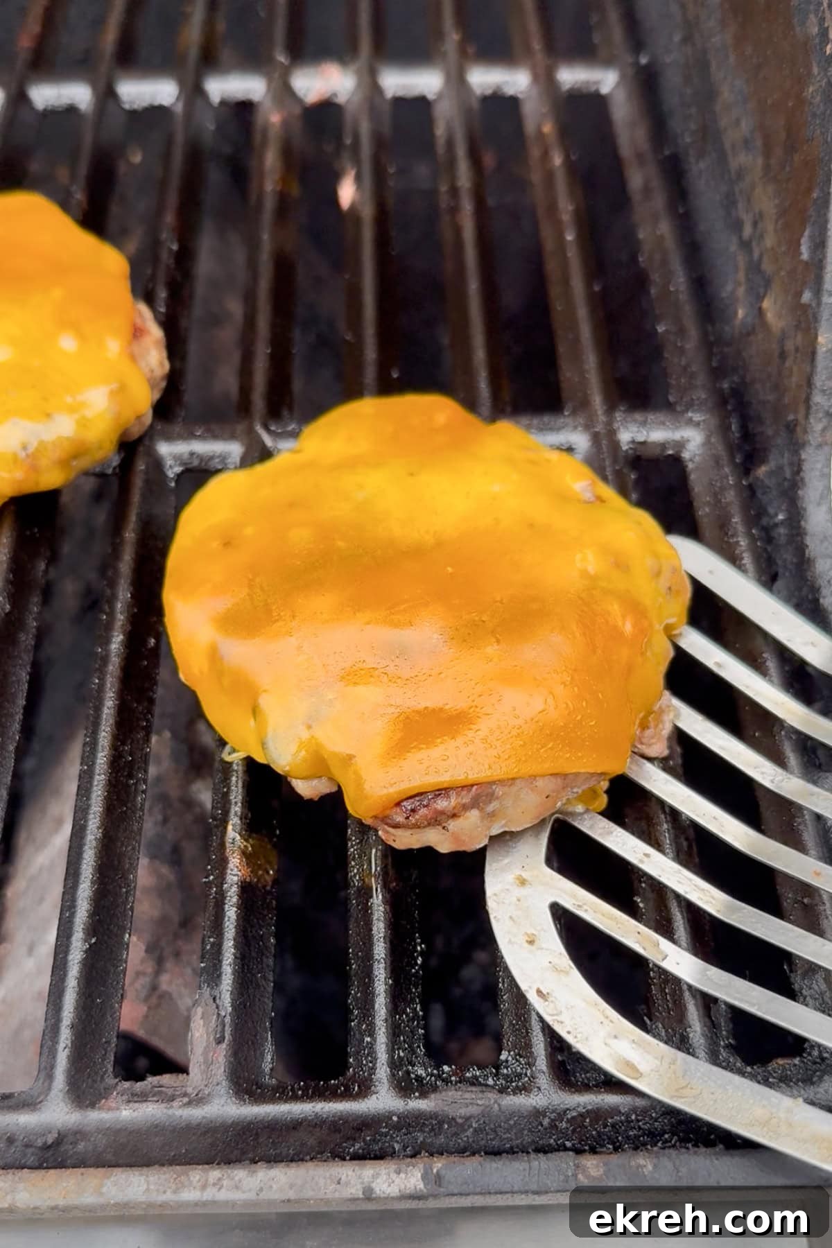 Juicy Grilled Bacon Cheeseburgers with Beef and Pork Patties 15 A close-up of a cheeseburger patty with melted cheddar cheese on a grill, with a metal spatula lifting the patty. Another patty is partially visible on the left.