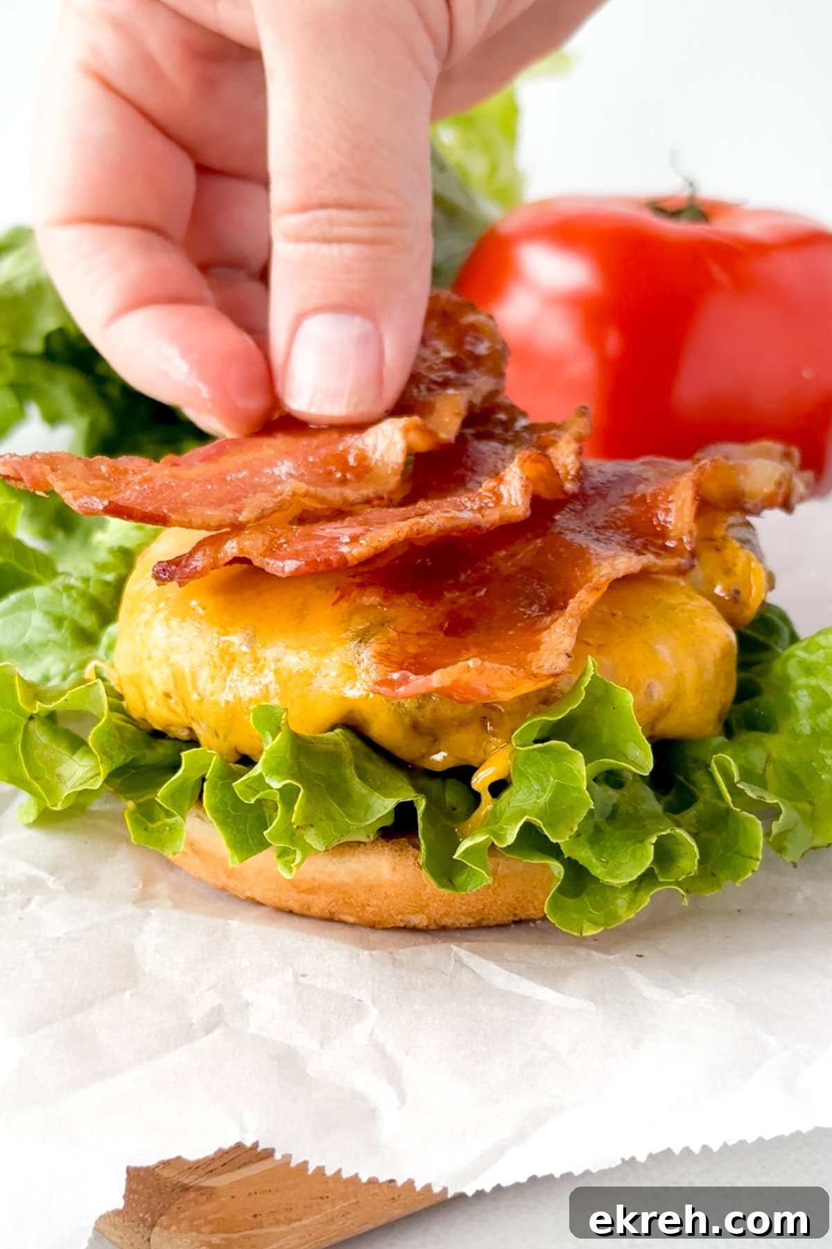 Juicy Grilled Bacon Cheeseburgers with Beef and Pork Patties 17 A hand places crispy bacon strips on top of a cheeseburger with fresh green lettuce on a hamburger bun. A whole tomato and leafy lettuce are visible in the background.