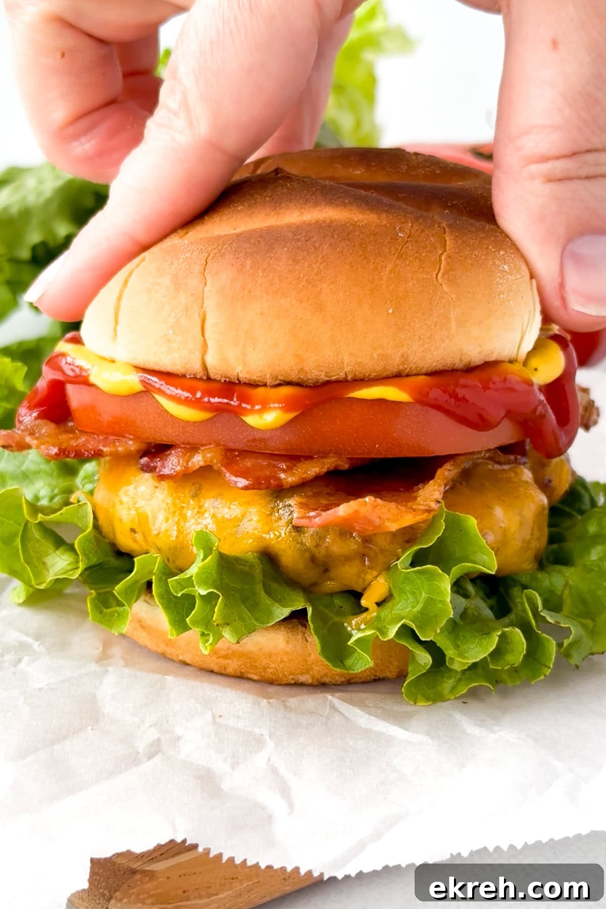 Juicy Grilled Bacon Cheeseburgers with Beef and Pork Patties 18 A hand holding the top bun of a cheeseburger with lettuce, tomato, bacon, cheddar cheese, and ketchup, set on white parchment paper with fresh lettuce in the background.