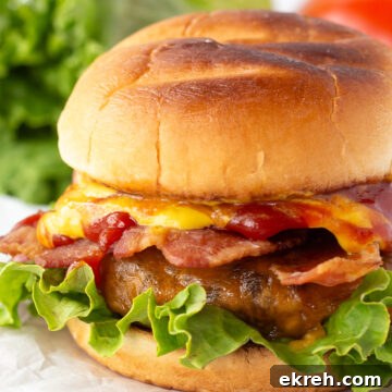A close-up of a hamburger with lettuce, bacon, melted cheese, and ketchup in a toasted bun, placed on a piece of parchment paper with lettuce and a tomato in the blurred background.
