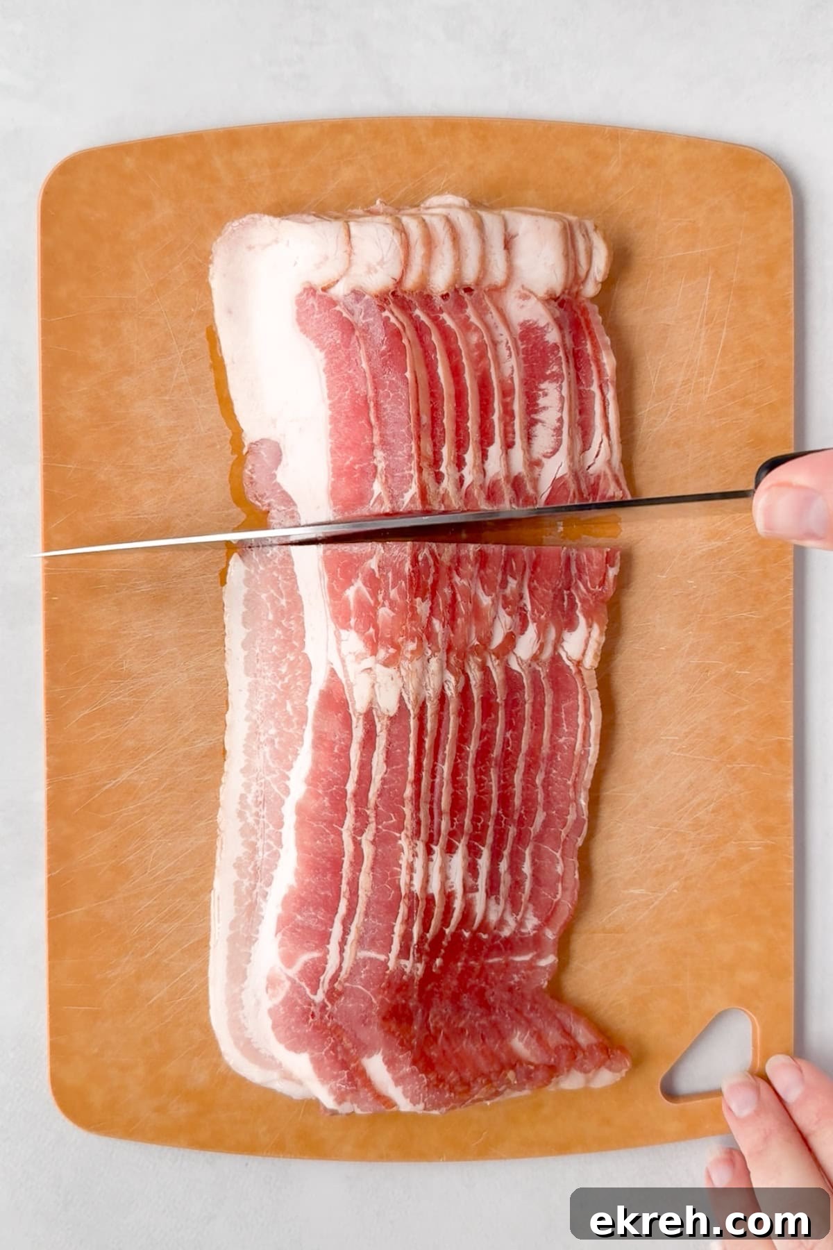 Juicy Grilled Bacon Cheeseburgers with Beef and Pork Patties 4 Several strips of raw bacon are stacked on a brown cutting board. A person is using a large knife to cut the bacon in half. The cutting board has a small triangular hole in one corner.