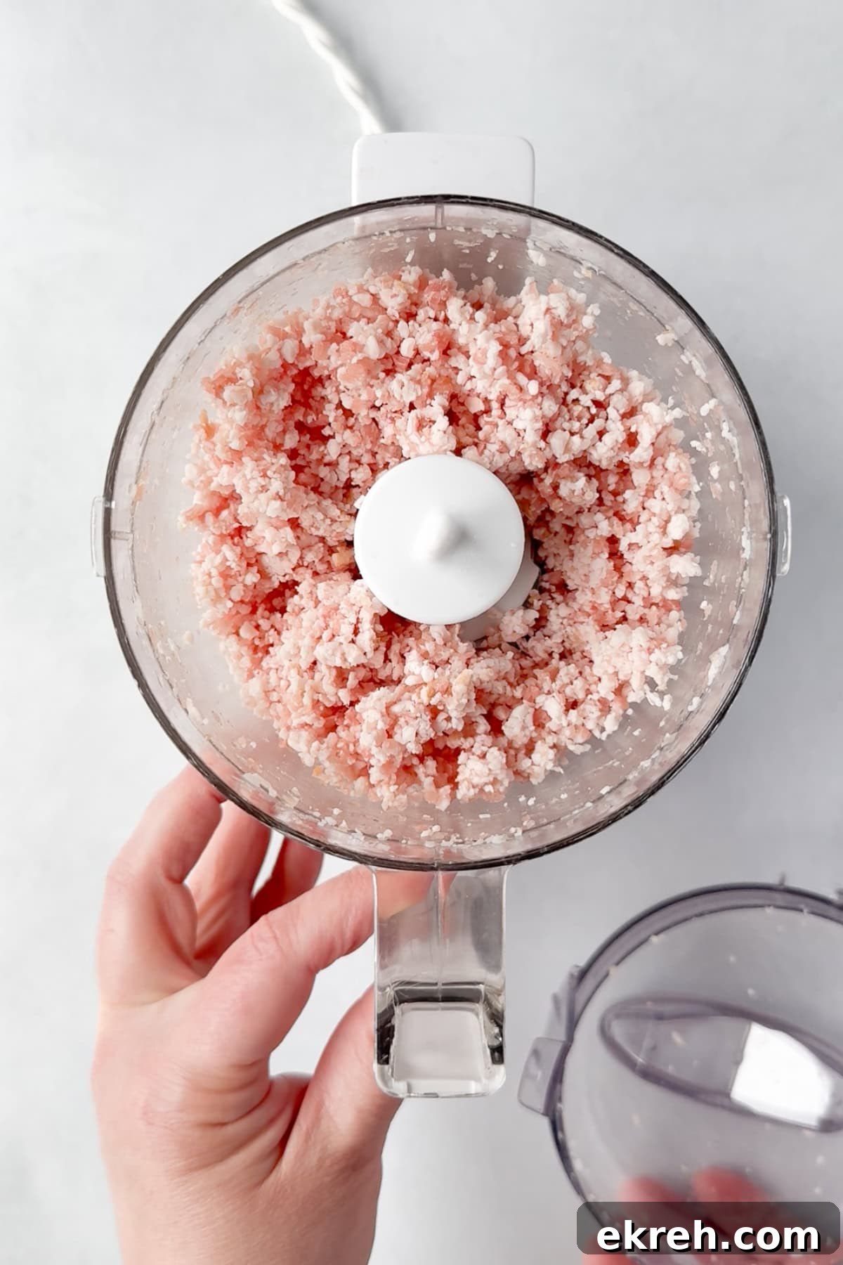Juicy Grilled Bacon Cheeseburgers with Beef and Pork Patties 6 A hand holds the base of a food processor filled with finely ground raw bacon, viewed from above on a white surface. The lid of the processor is removed and held in the other hand.