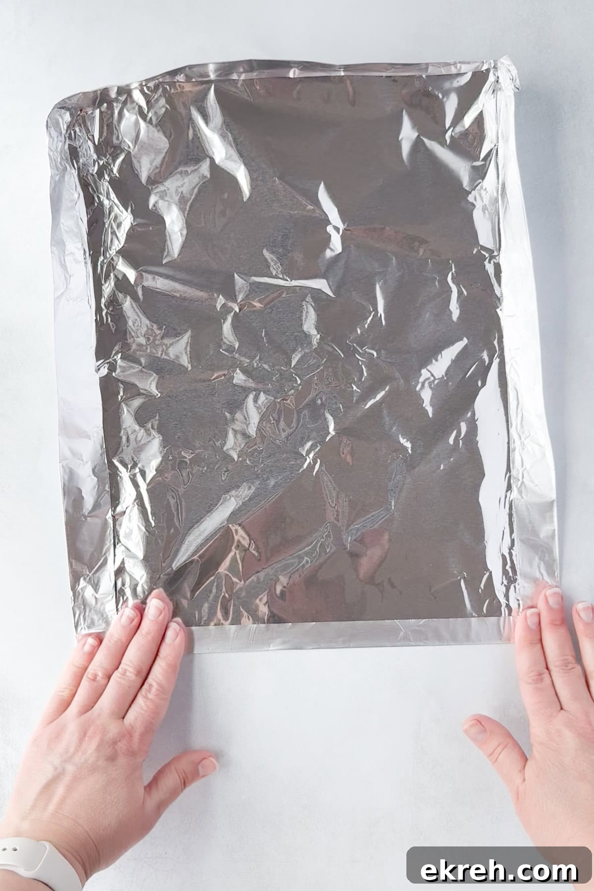 Juicy Grilled Bacon Cheeseburgers with Beef and Pork Patties 7 Two hands are pressing down the edges of a sheet of aluminum foil, placed on a white surface.