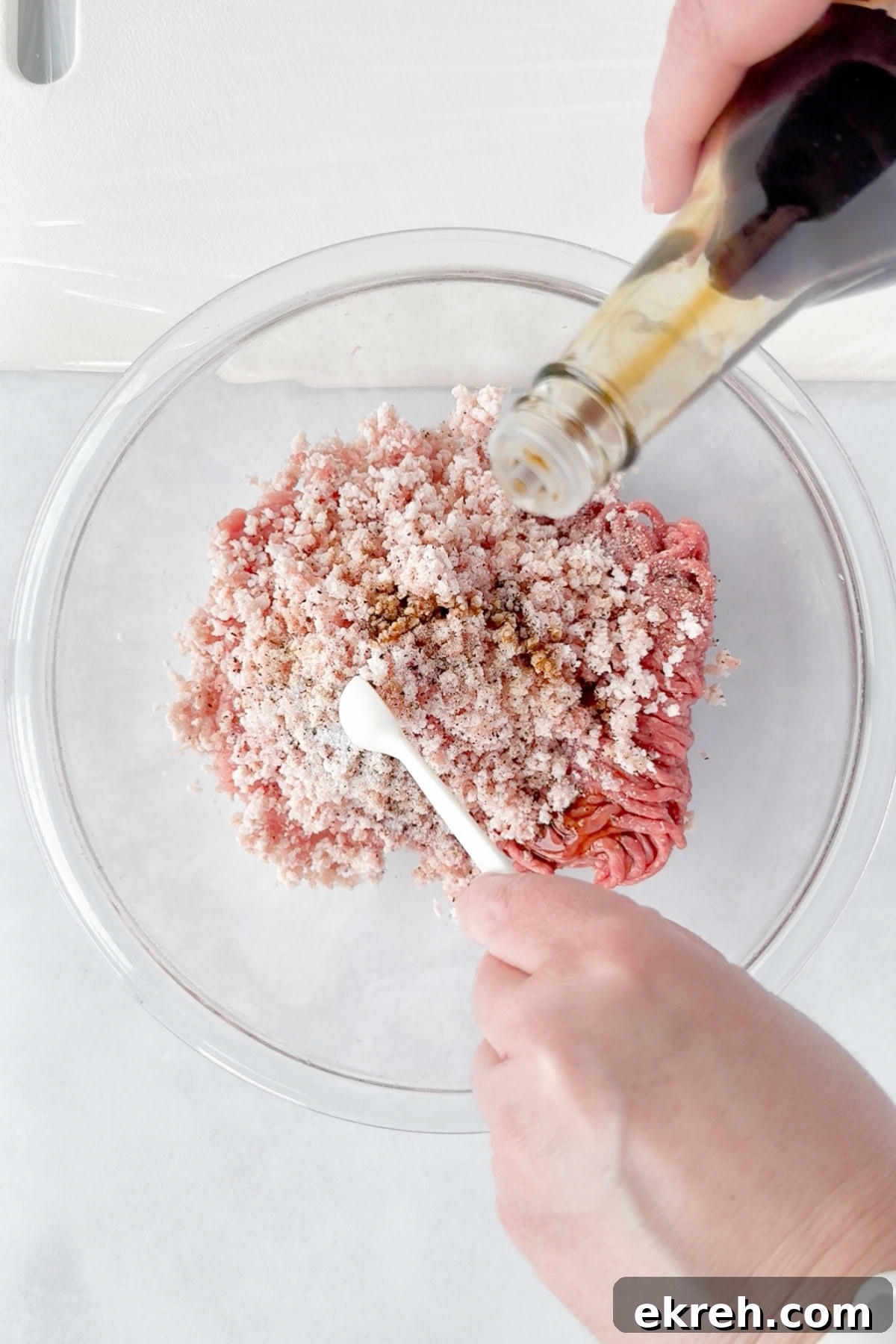 Juicy Grilled Bacon Cheeseburgers with Beef and Pork Patties 8 A person pours liquid seasoning from a bottle into a glass bowl filled with ground meat and seasonings, while holding a little white measuring spoon.