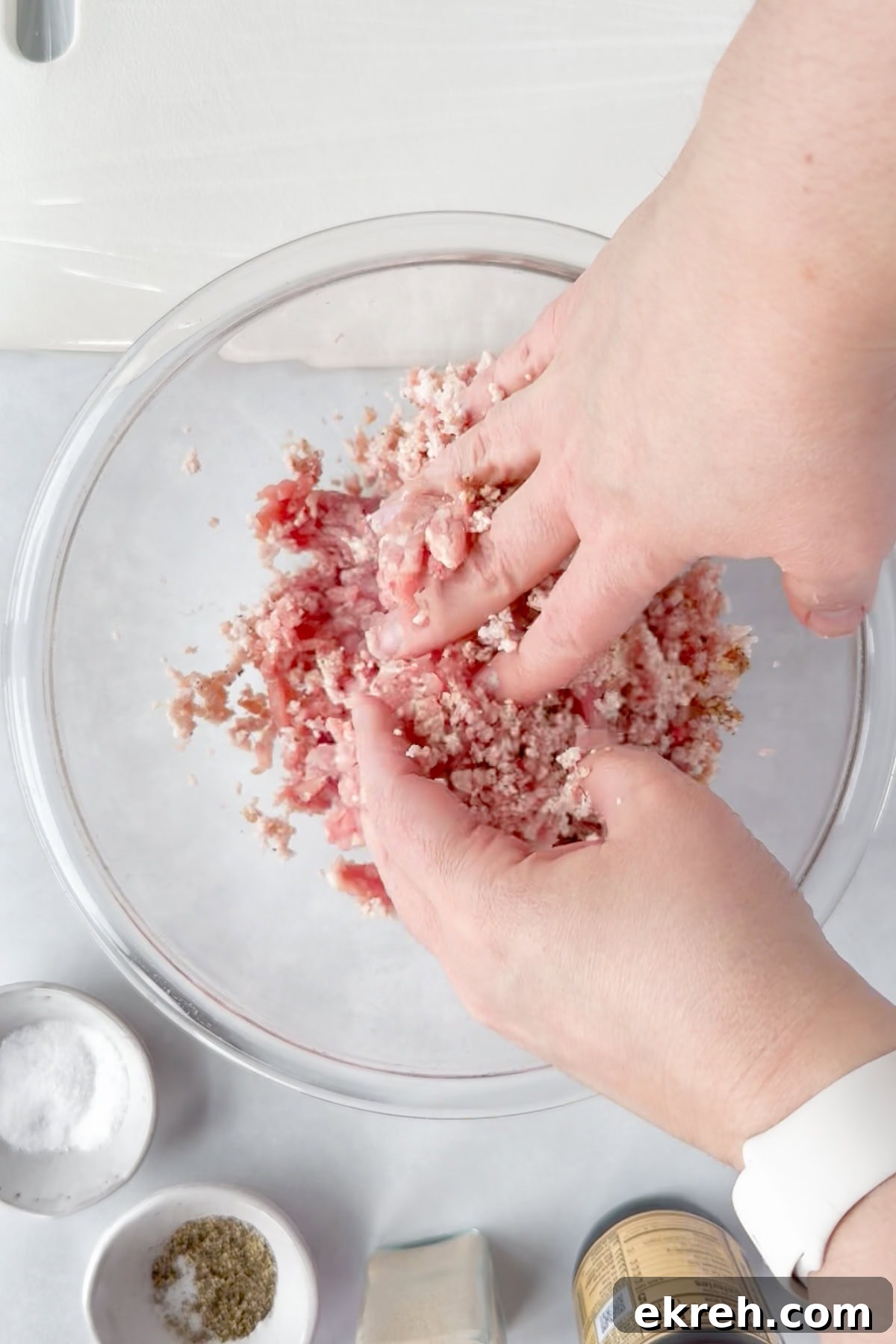 Juicy Grilled Bacon Cheeseburgers with Beef and Pork Patties 9 Two hands mix ground meat with seasoning in a clear glass bowl. Surrounding the bowl are small dishes containing salt, herbs, and a jar of seasoning on a white countertop.