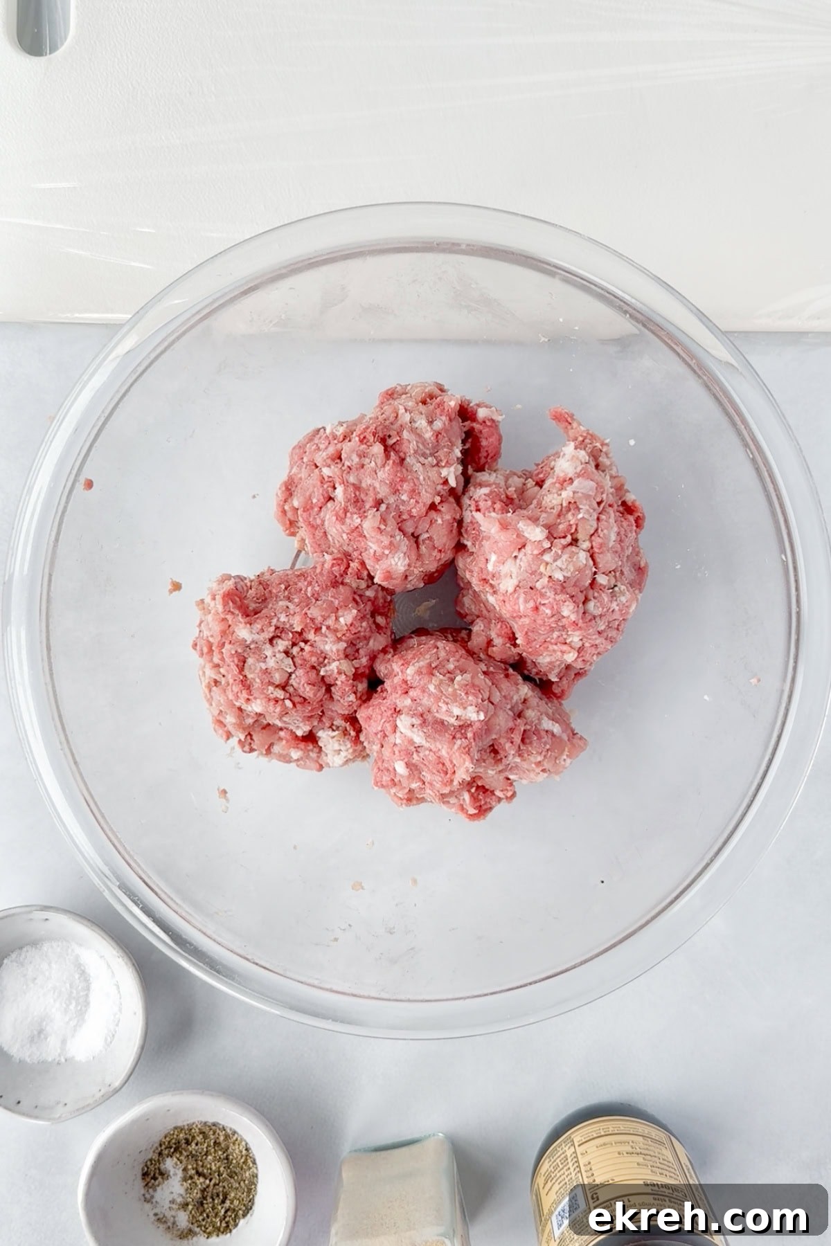 Juicy Grilled Bacon Cheeseburgers with Beef and Pork Patties 10 A clear glass bowl containing four portions of raw ground meat sits on a white surface. Surrounding the bowl are small dishes with salt, dried herbs, a jar of seasoning, and a spice container.