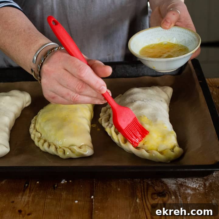How to Make a Traditional Cornish Pasty (Authentic Recipe) 15 womans hands brushing beaten eggs with a red silicone brush onto three uncooked Cornish pastys on a black baking tray
