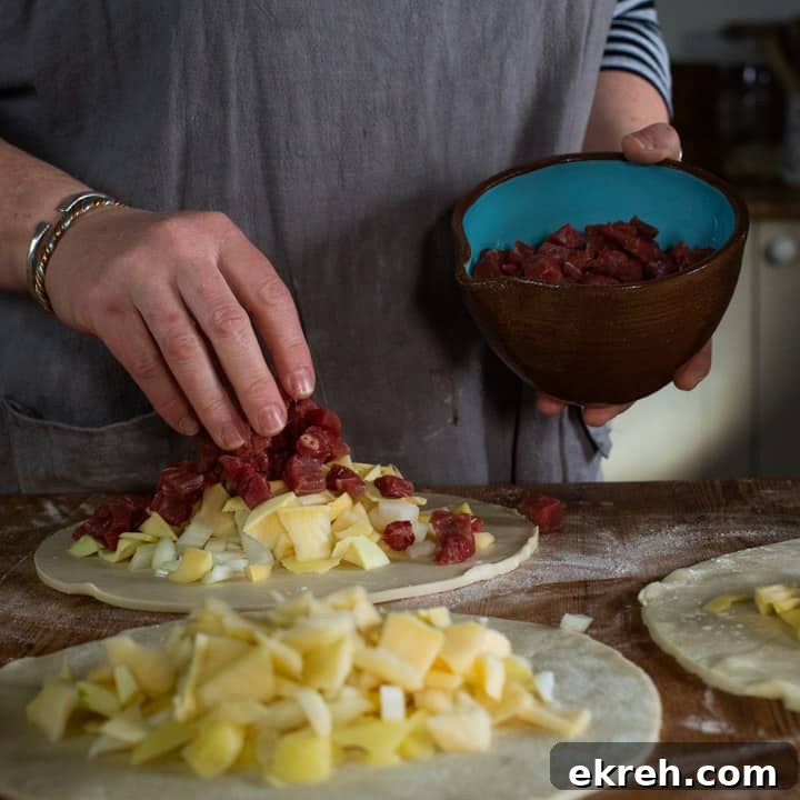 How to Make a Traditional Cornish Pasty (Authentic Recipe) 9 three discs of pastry on a wooden surface, piled up with raw swede, potato, onion and steak ready to be crimped into pasty's