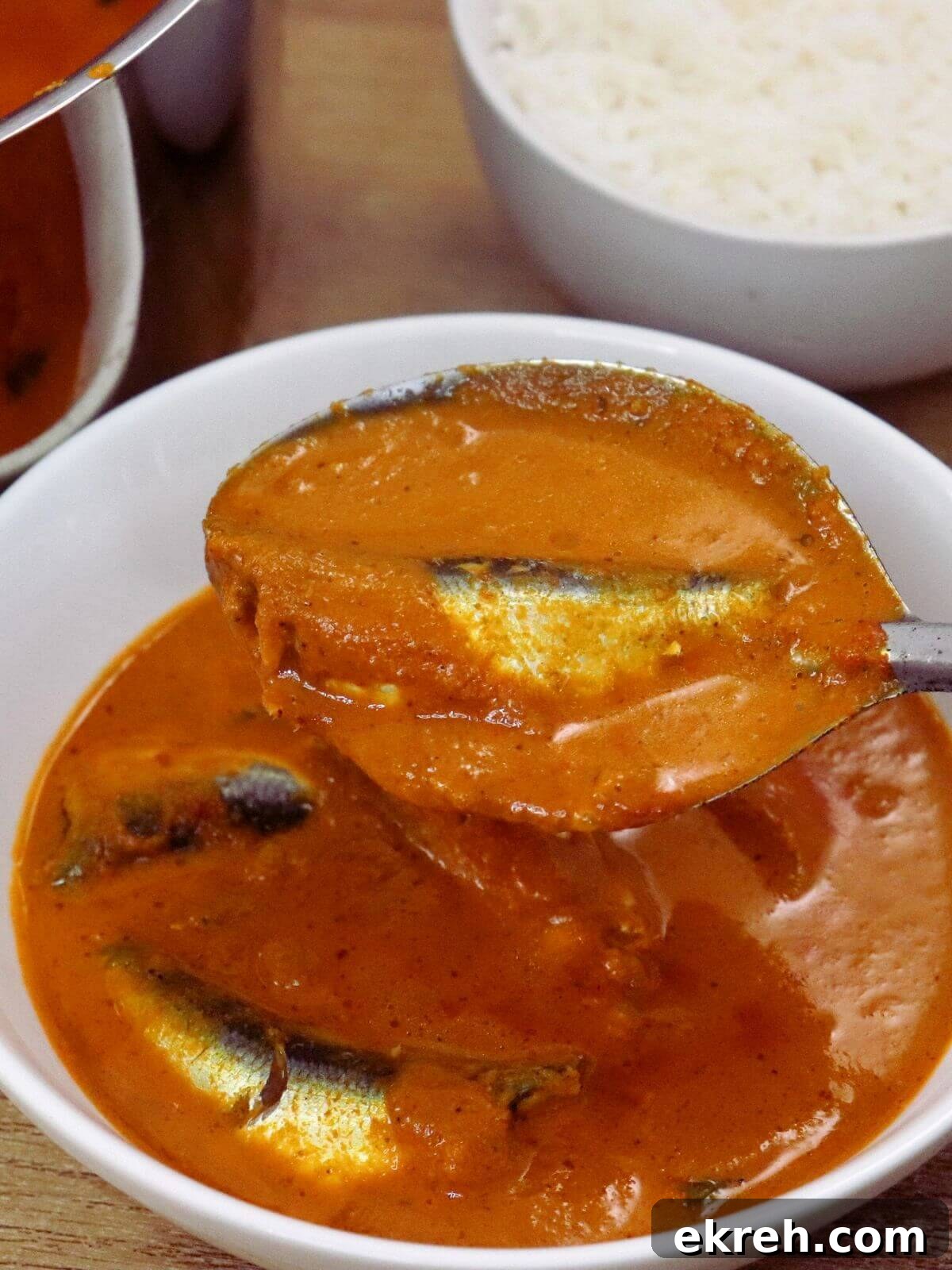 A close-up view of a bowl of sardine curry, with a ladle full of curry held above it.