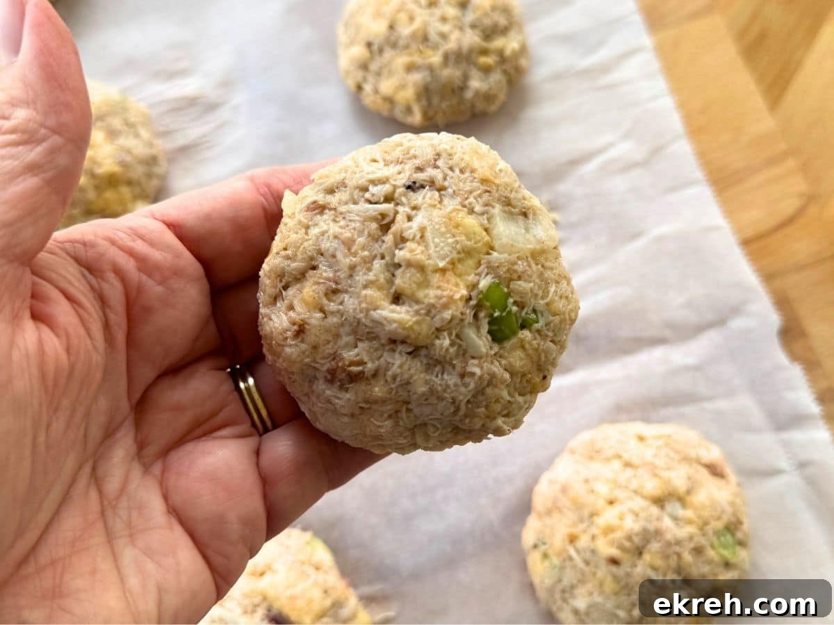 Hand holding an uncooked Maryland-style lump crab cake with a slightly domed shape over parchment paper with more crab cakes on it.