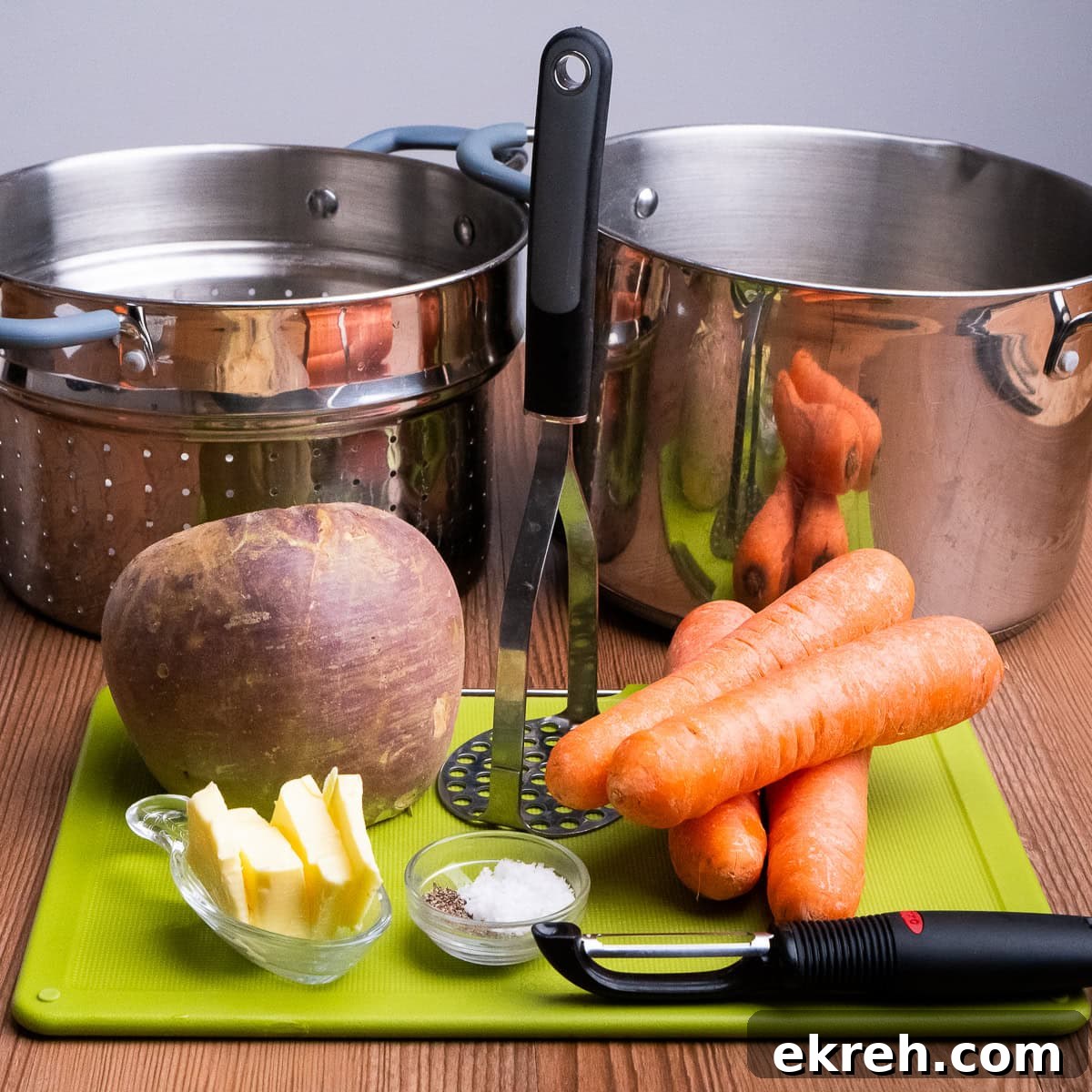 Four carrots,one swede,butter, salt pepper in glass dishes, a peeler, a masher, a green chopping board, a large cooking pot and sieve
