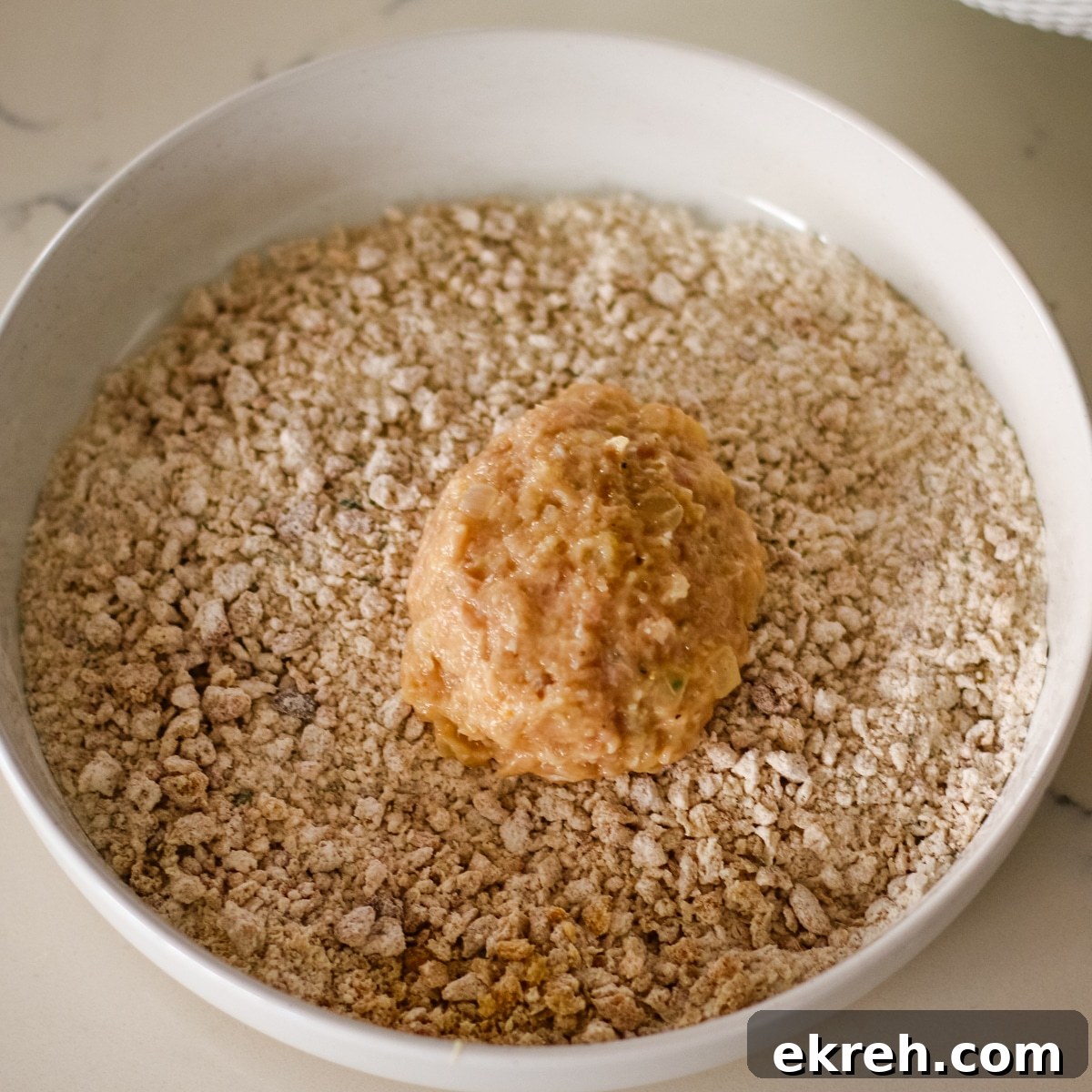 raw, round chicken mixture patty being coated in coarse breadcrumbs inside a white bowl.