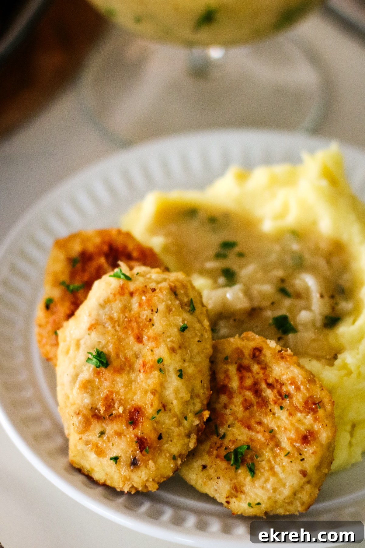 three golden-brown chicken kotleti served on a white plate with a side of mashed potatoes topped with gravy.