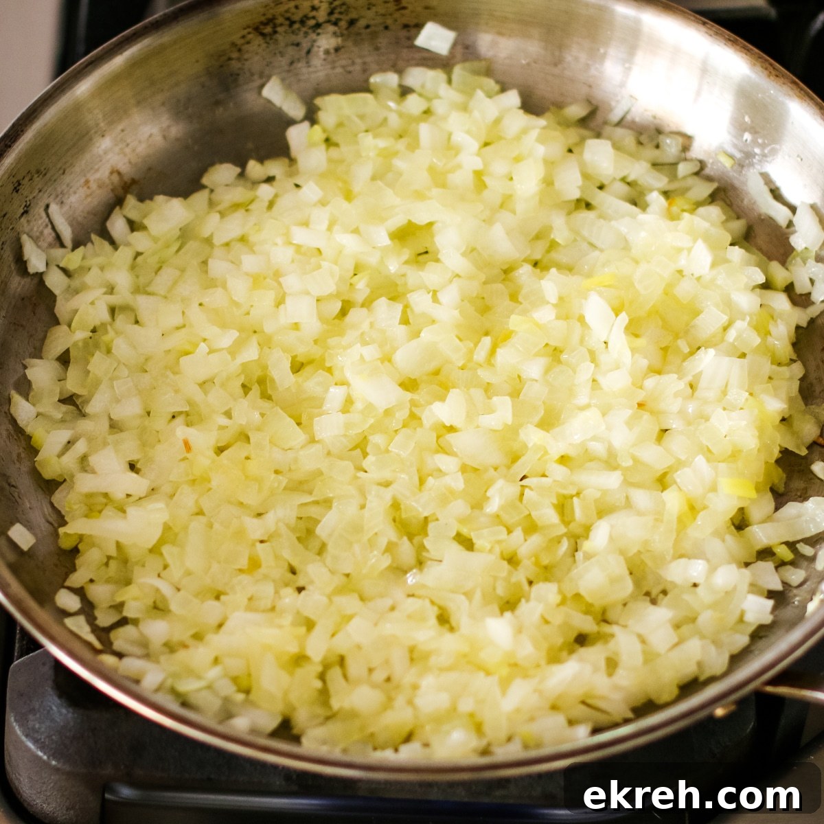 large amount of finely chopped white onions sautéing in a stainless steel skillet on a stovetop.