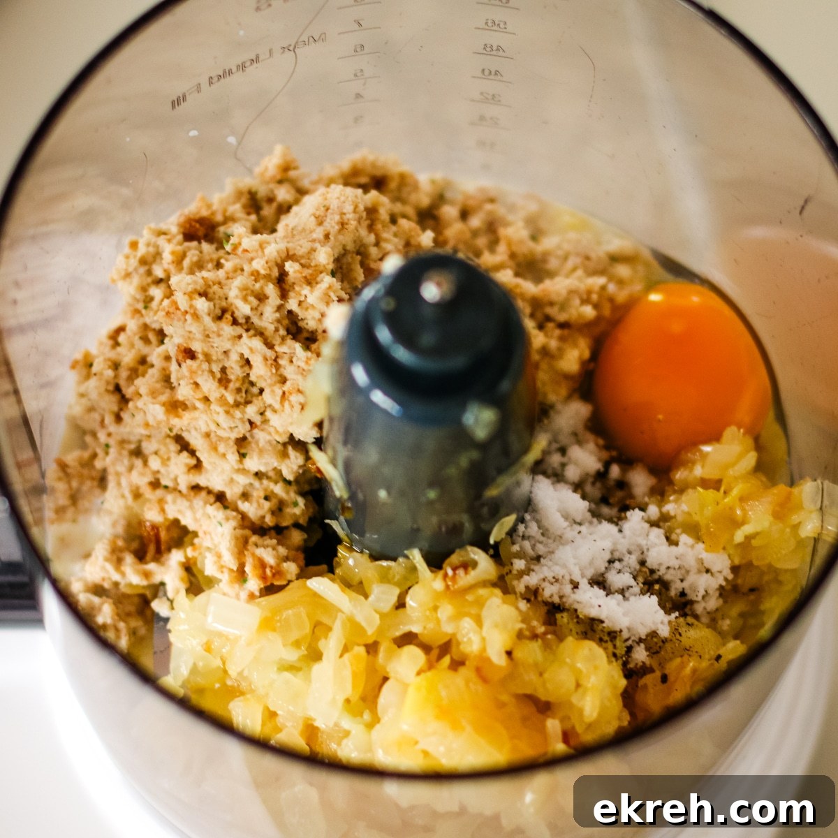 food processor bowl containing ground bread and meat mixture, chopped sautéed onions, an egg yolk, and a pile of salt, ready to be blended.