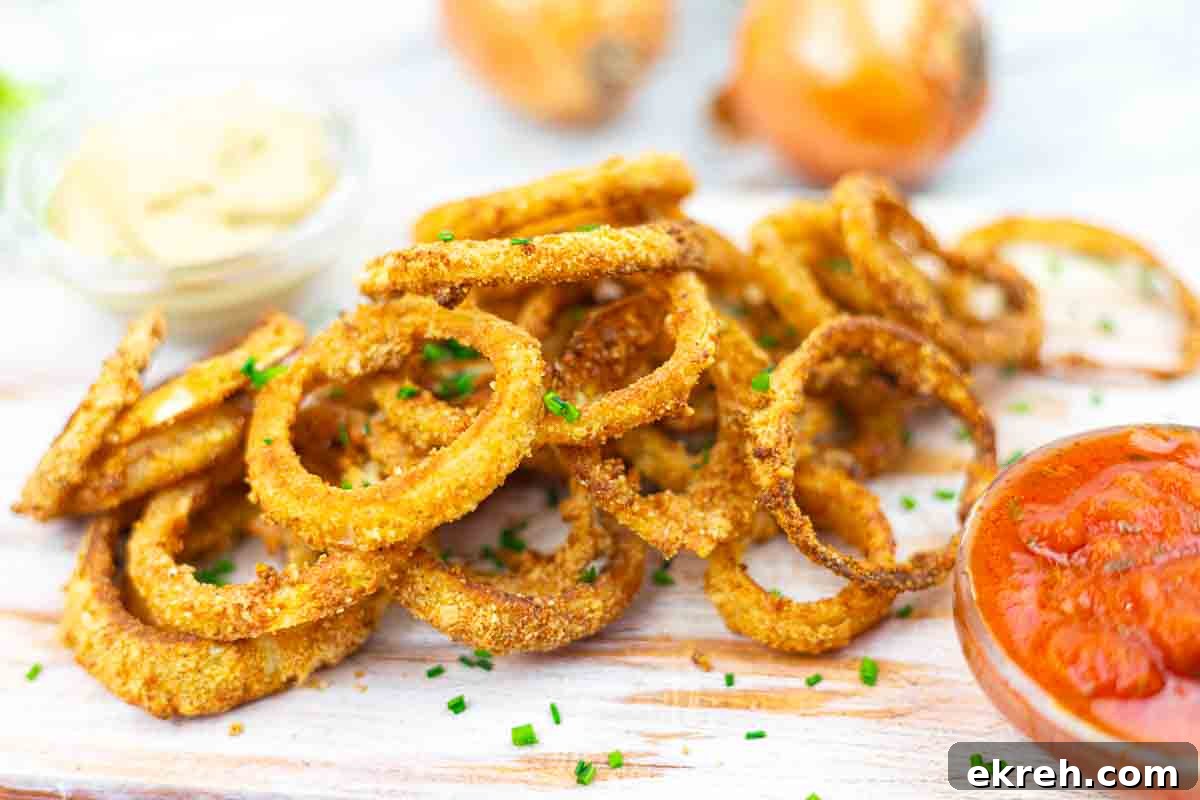 A pile of crispy onion rings garnished with chives, accompanied by small dishes of dipping sauces, are shown on a light wooden surface.