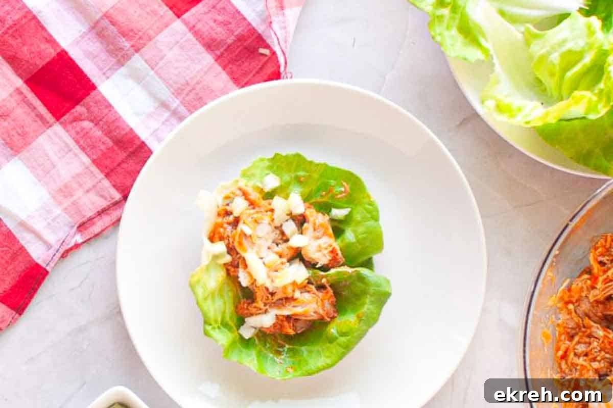 A white plate with a lettuce leaf topped with shredded chicken and diced onions, next to a red and white checkered cloth and fresh lettuce leaves.